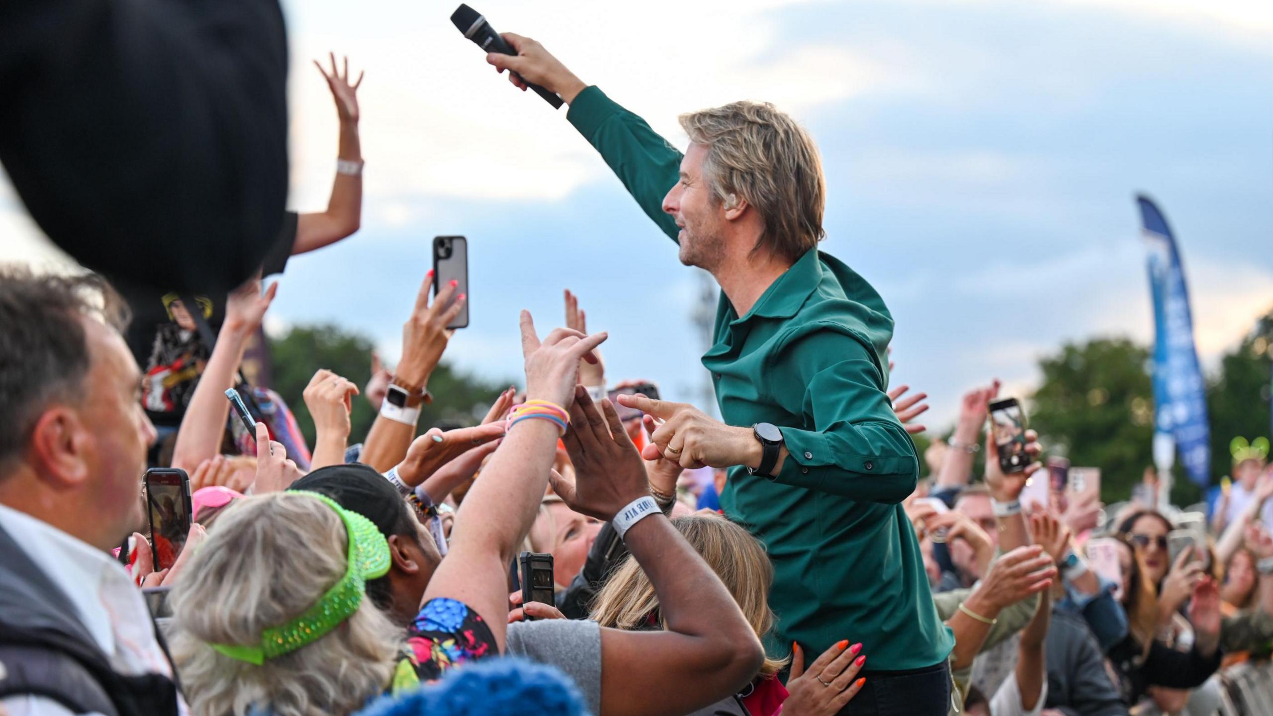 Singer Chesney Hawkes, with blonde hair and a green shirt, leans over a barrier into a crowd of people dancing to his music, holding a microphone aloft for them to sing into