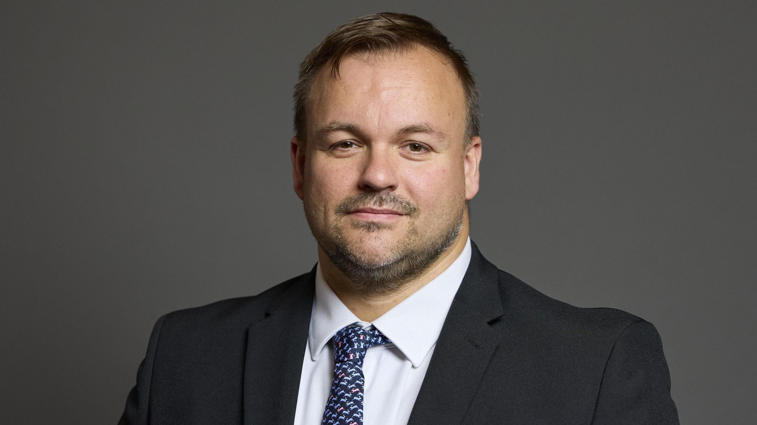 Terry Jermy is wearing a black suit with a white shirt and a blue tie. 
He has short, brown hair and a short beard and moustache.
He is standing in front of a grey background for his official portrait from the House of Commons.