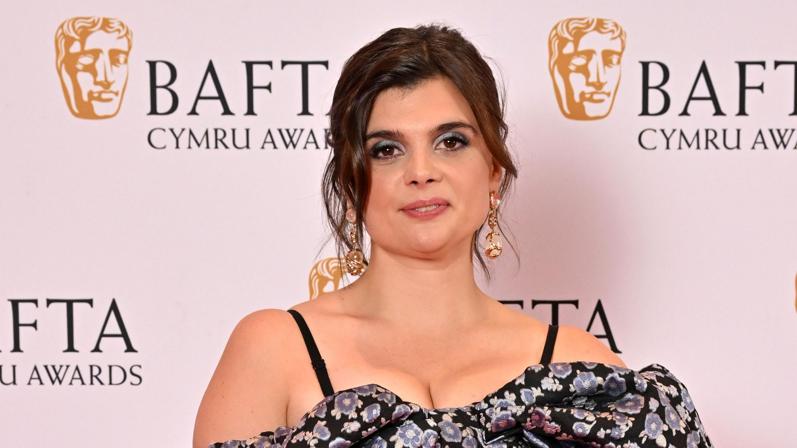 A woman with brown hair, dark eye makeup, gold earrings, and a blue and dark floral dress poses for a photo on the Bafta Cymru red carpet, with Bafta images on a white wall behind her.