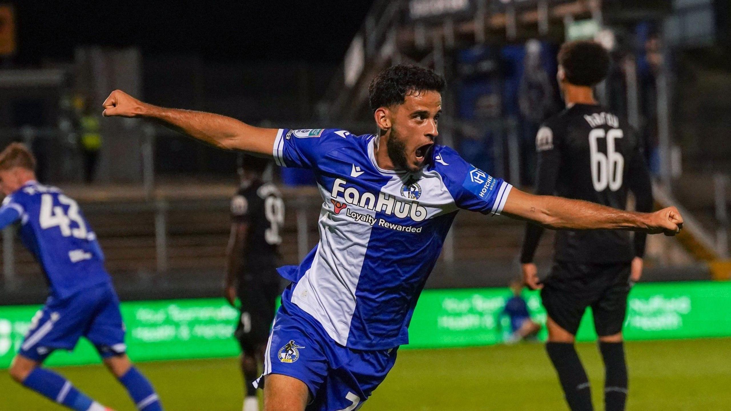 Fabrizio Cavegn celebrates a goal for Bristol Rovers against Tottenham Hotspur Under-21s in the EFL Trophy. He has his arms outstretched and his mouth wide open. He is wearing the traditional Rovers kit of blue and white quarters.