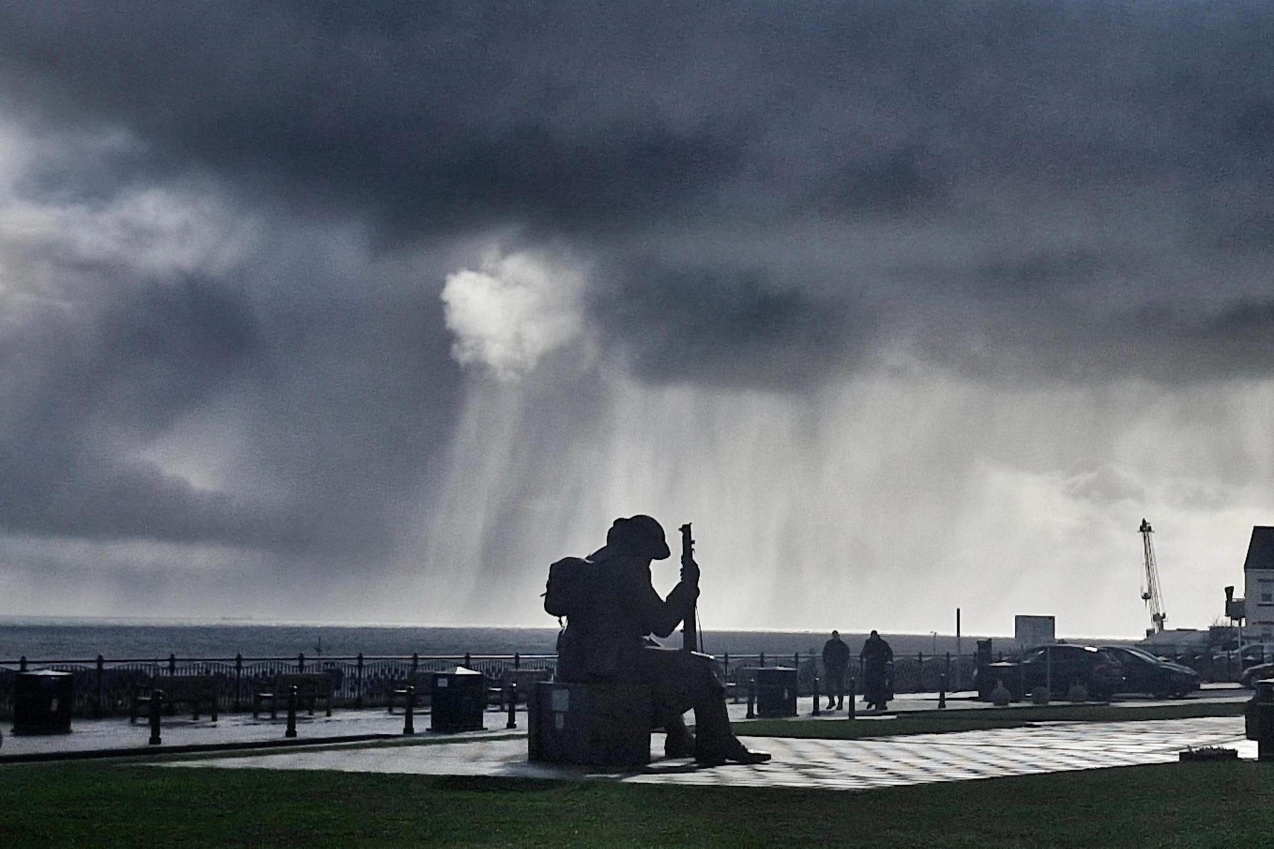 A statue of a man in military gear with his head bowed is illuminated in silhouette. Rain clouds are overhead.