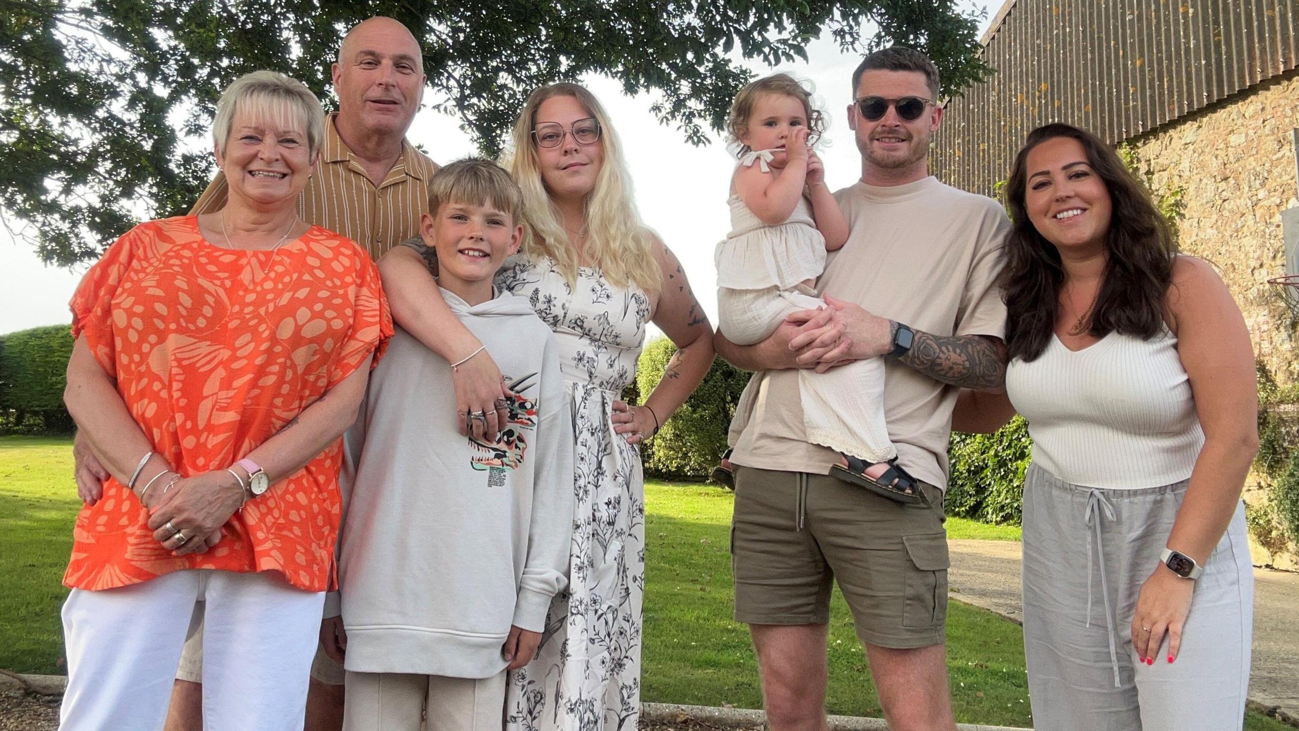 The Roberts family, standing outside in summer clothing looking at the camera. Pictured are Angela and Martin, their daughters Nikki and Becky, Nikki's partner Liam and their children Fin and Cleo.