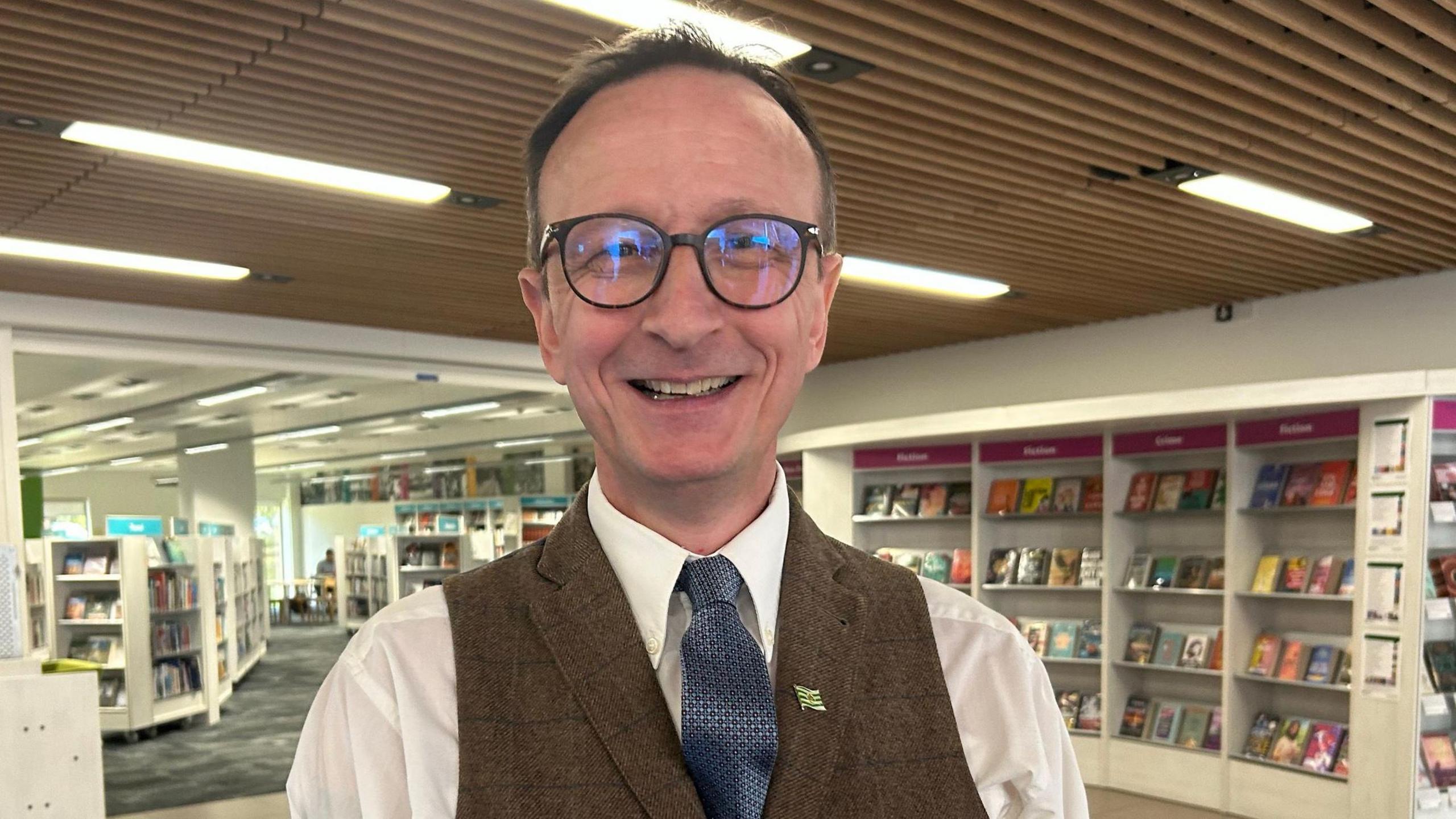 Ian Hicks smiling at the camera wearing glasses, white shirt & tie and a tweed waistcoat. He is in Trowbridge Library.