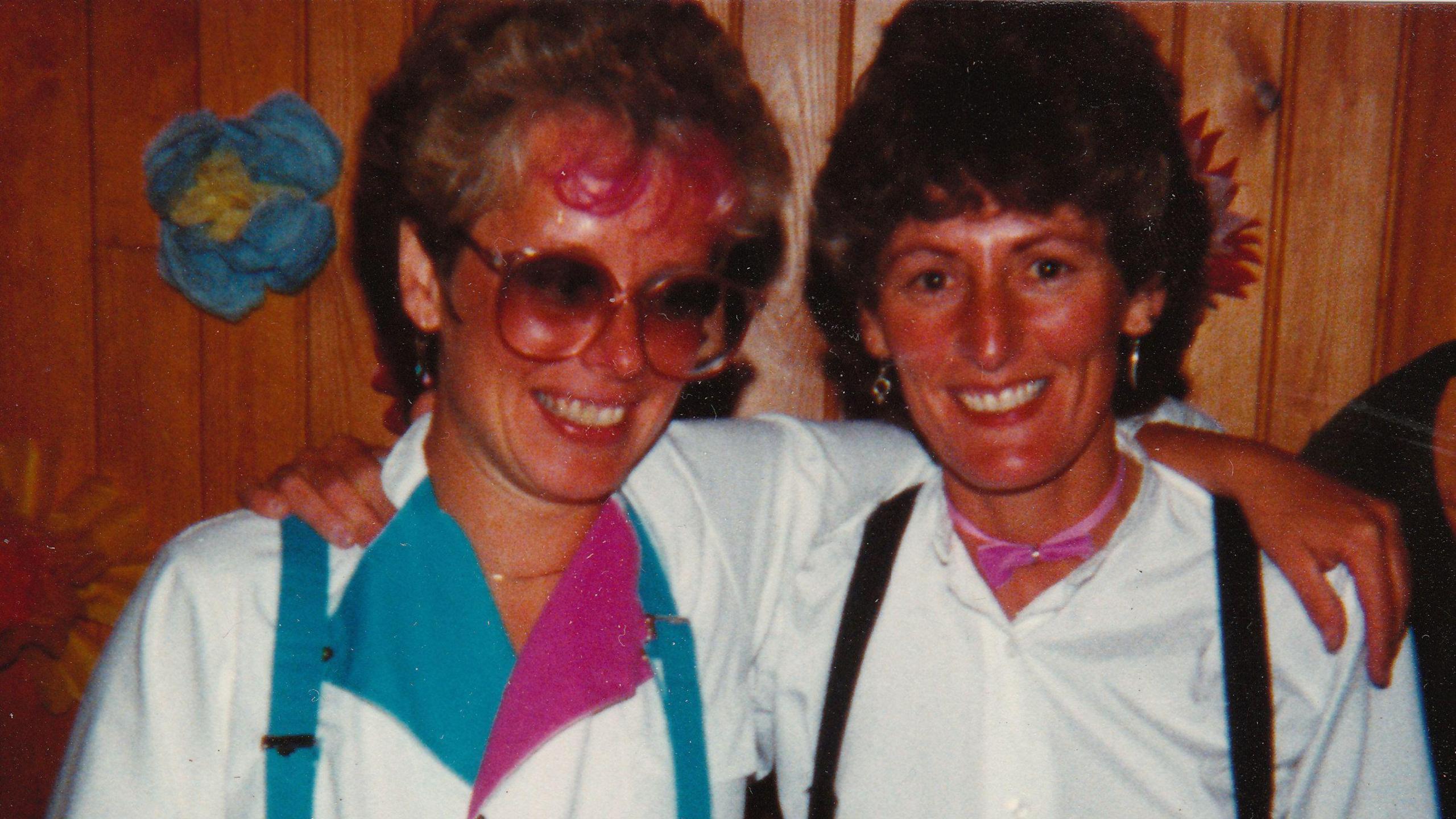 Anita and Steph in 1981, in a colour photo, Anita has on large glasses and a white top with blue and pink on, and blue braces, Steph has a white top on with black braces, they are both standing in front of a wooden panelled wall. 