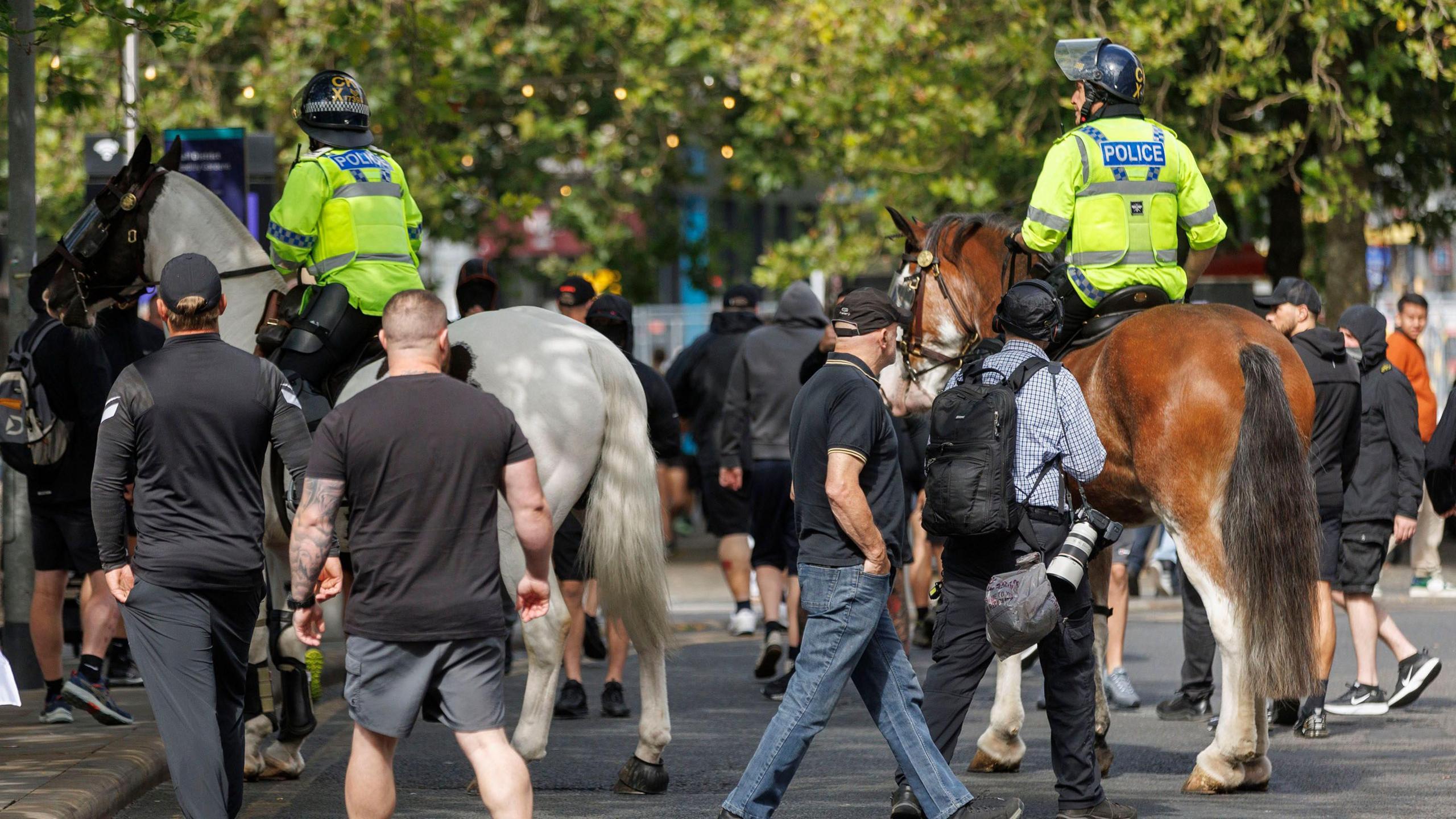 Members of the public walk behind mounted officers on two horses on a street in Manchester. The men walking behind are wearing dark clothing.