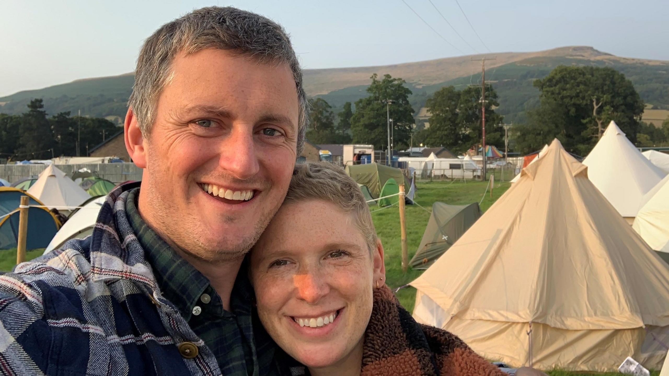 Phillippa and Matt Hentsch at a music festival in a field, with tents and hills in the background