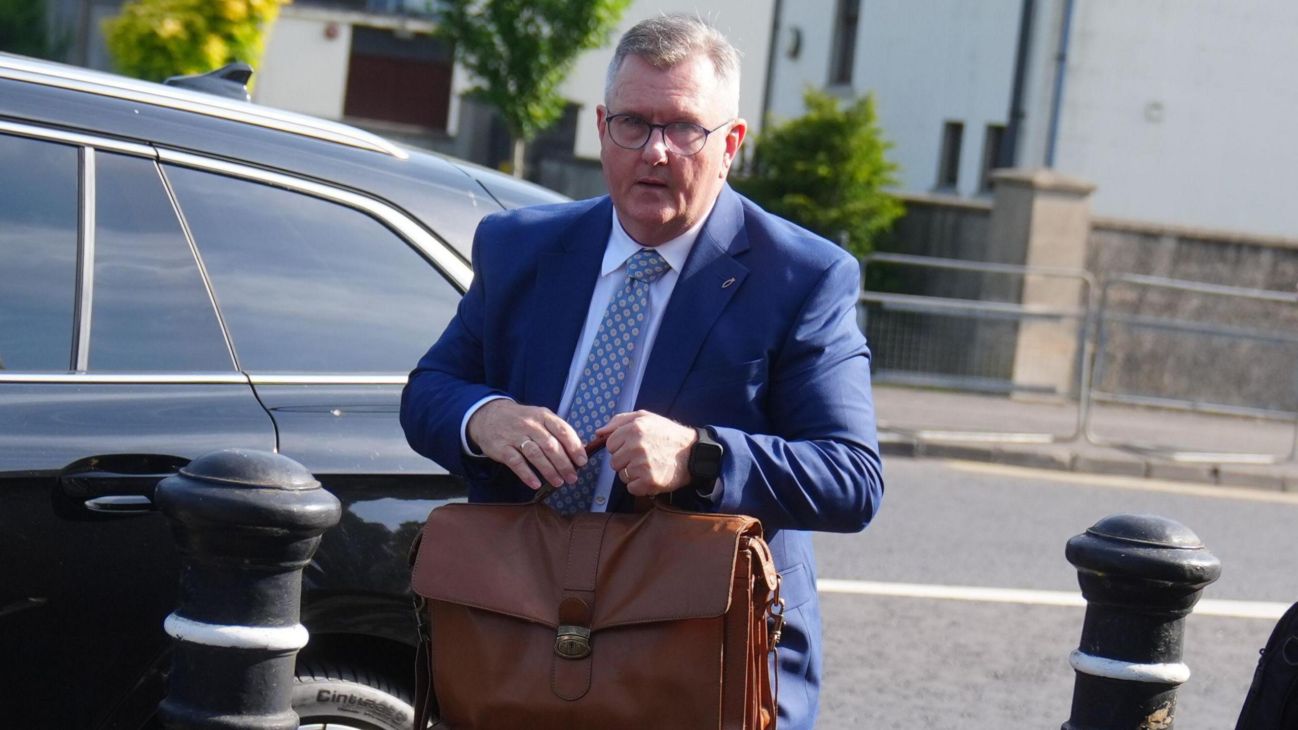 Sir Jeffrey Donaldson has short, cropped grey hair. He is wearing black-rimmed glasses and a blue suit, white shirt and blue patterned tie. He is standing in front of a black car and is carrying a brown leather satchel bag in front of him. 