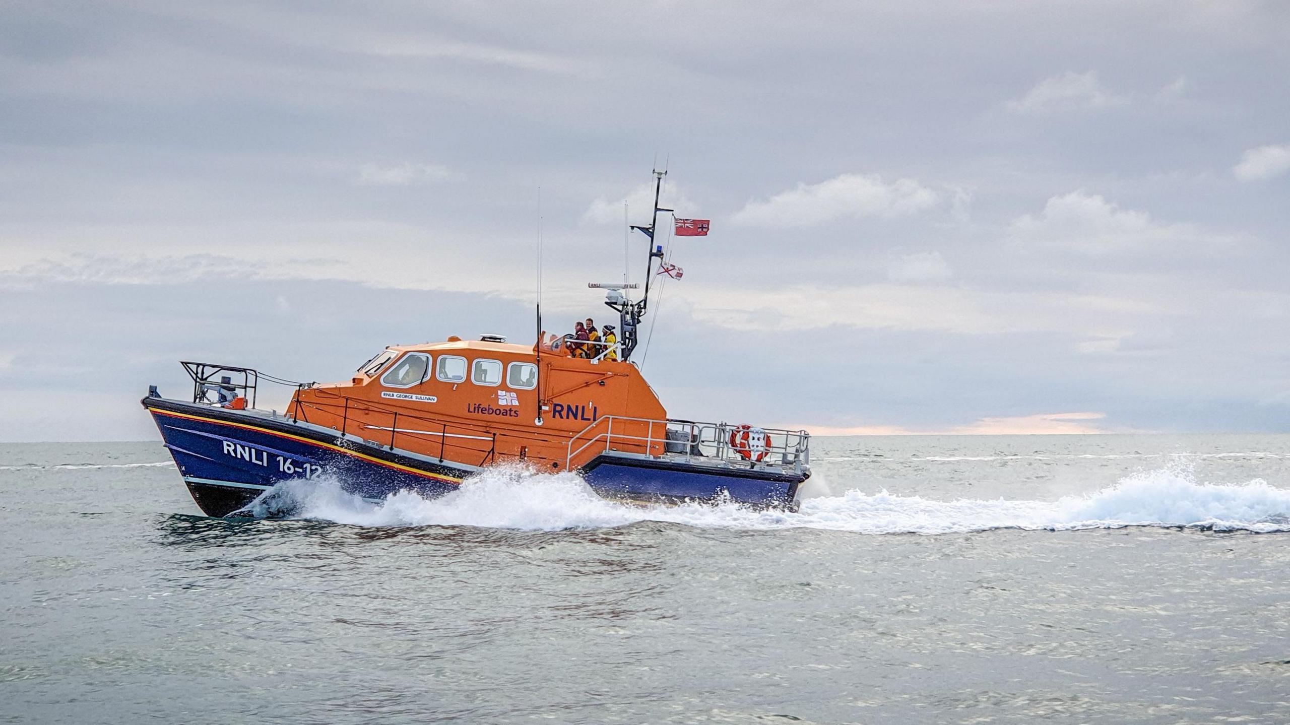 The RNLI lifeboat at sea. The ship is in motion with water splashing up the side of it. The ship is painted in two halves. The bottom is dark blue and the top is orange. A number of people are at the top of the boat where it is steered