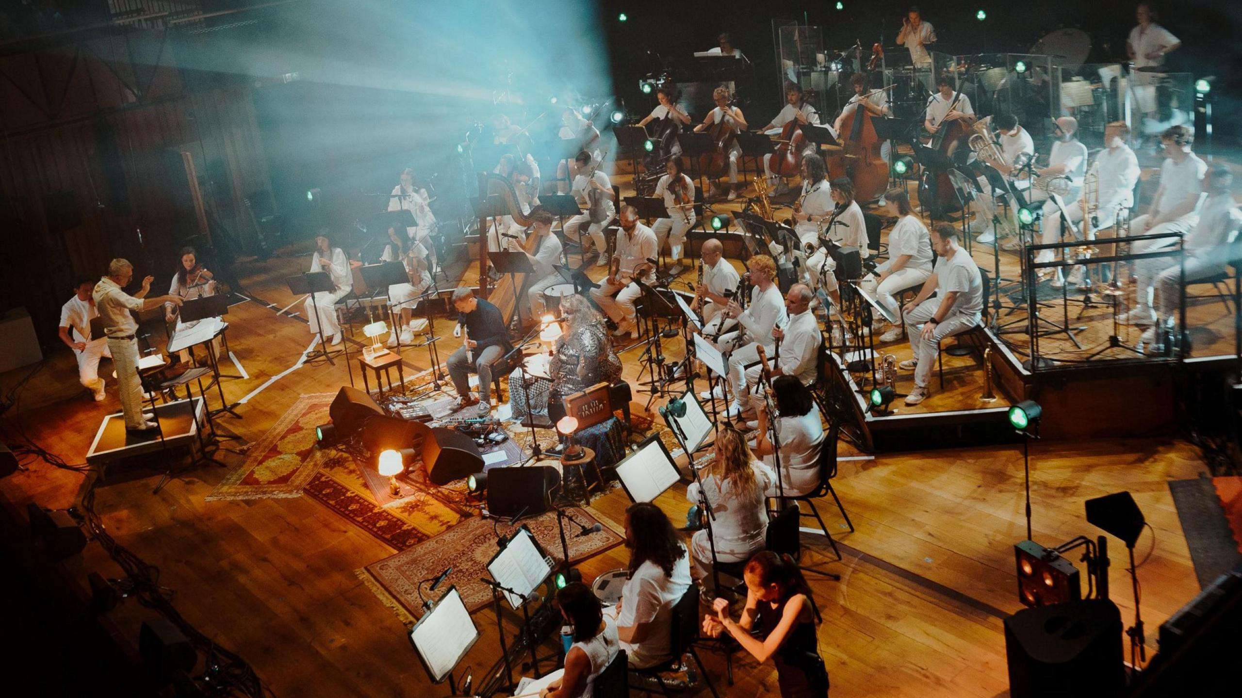 Members of the Paraorchestra are seen from the side stalls at the Bristol Beacon during the BBC Proms. They are all dressed in white and arranged in semi-circular rows. They are being conducted by a man who is also dressed in white.
