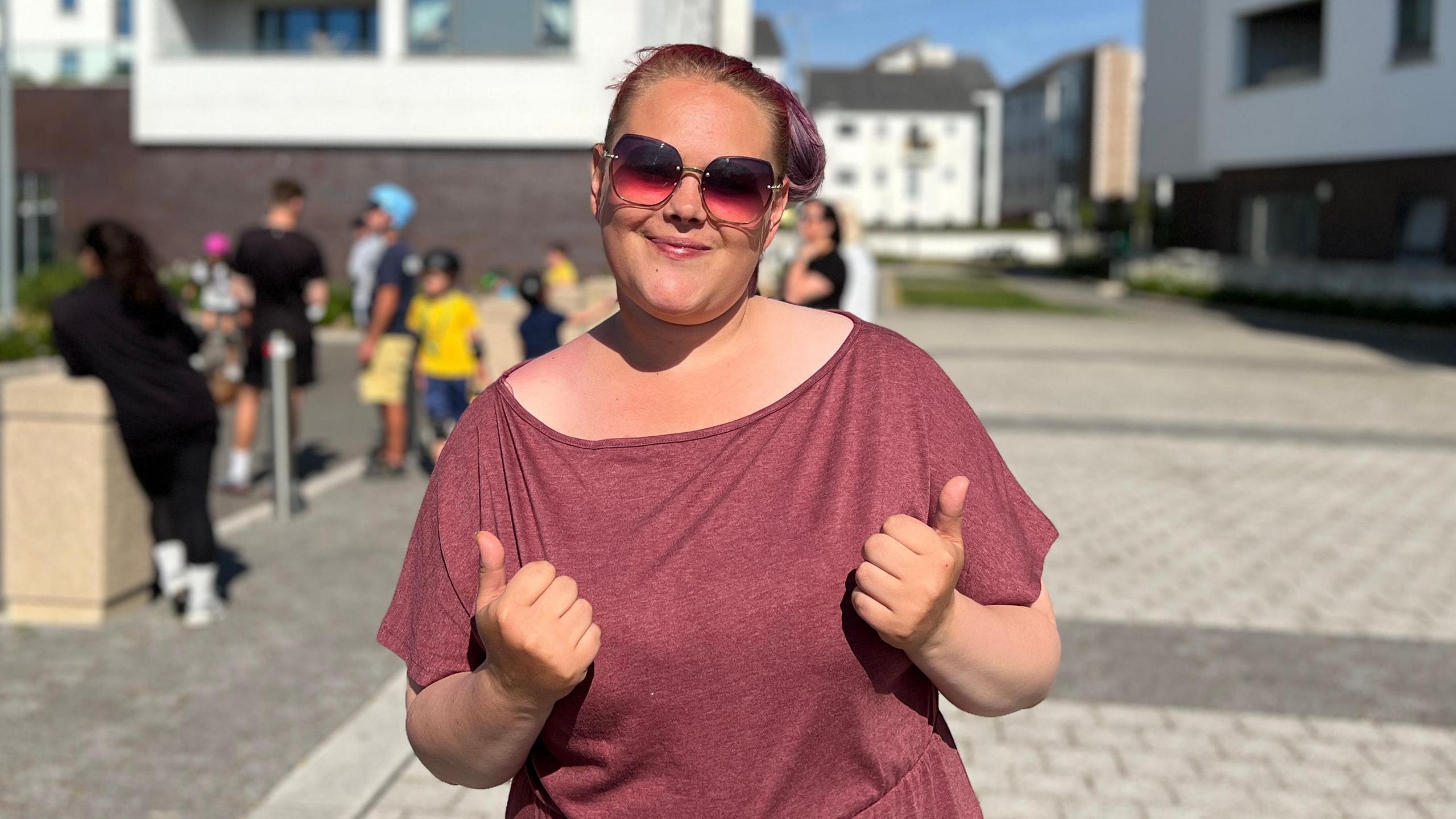 The image depicts a person standing in an outdoor urban setting with modern apartment buildings in the background. They are wearing a maroon dress and giving two thumbs up. The scene is lively, with several other people, including children, visible in the background, seemingly engaged in different activities. The weather appears pleasant, with a clear sky and some clouds, and the area is brightly lit by sunlight.