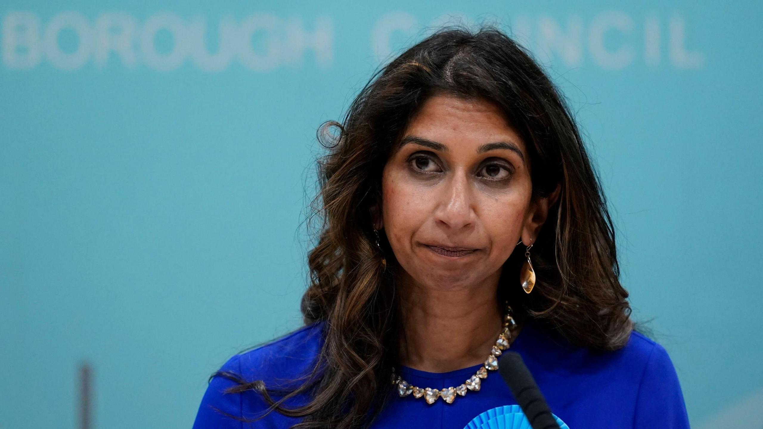 Former home secretary Suella Braverman in  blue dress speaking into a microphone. A light-blue wall is in the background