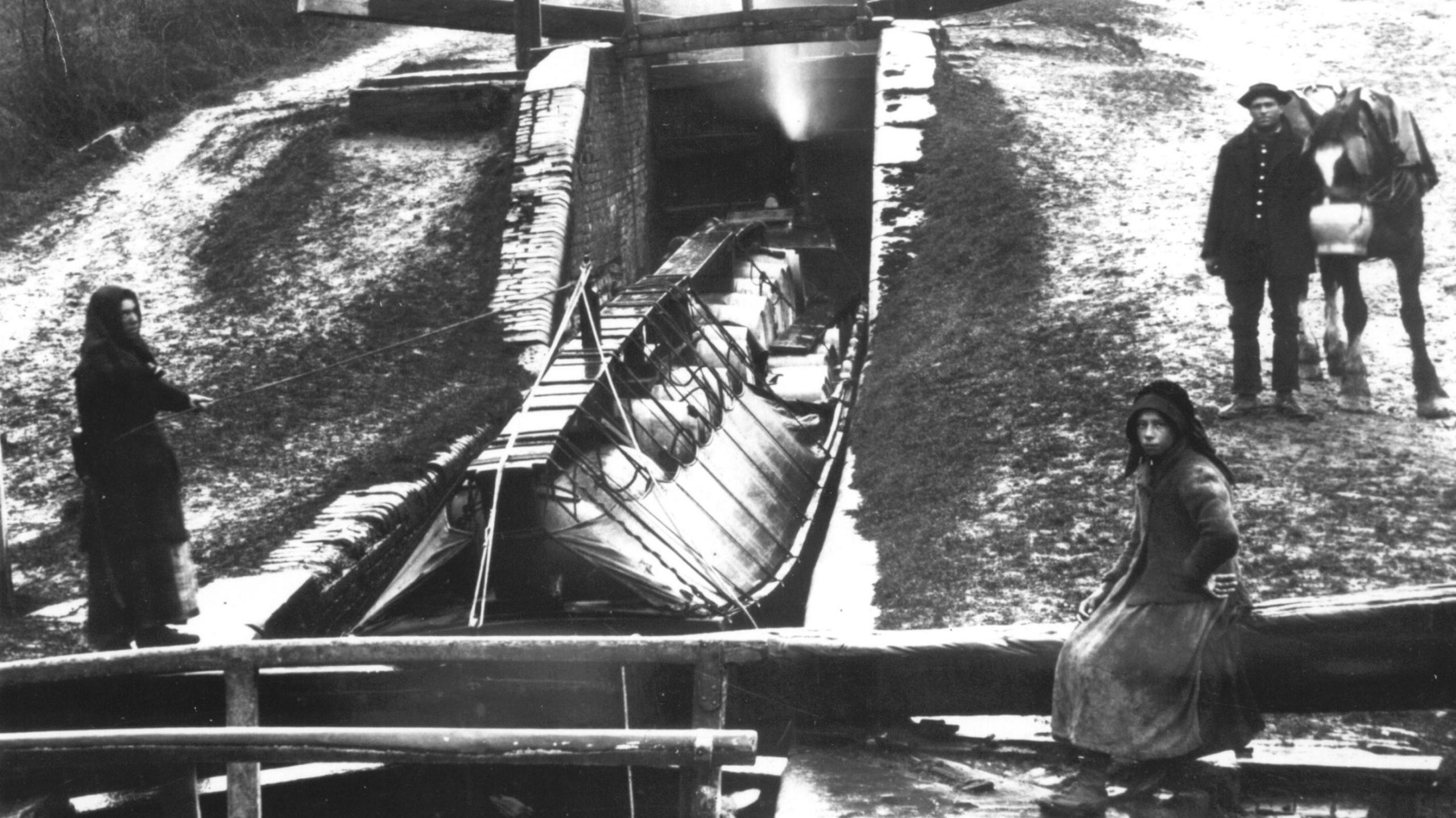 An old black and white image taken of Foxton Locks in Leicestershire on a snowy day. It shows canal boat descending down the staircase of locks. A woman is sitting on the lock on the right of the image looking toward the camera while a man and horse stand behind her. Another woman on the left of the lock is holding a rod connected to the boat.