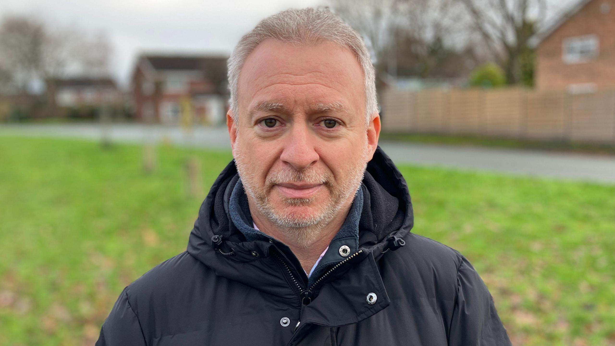 A grey-haired man posing for a photo wearing a black hooded coat on a residential road standing on a grassy bank. He has short blonde/white hair and a short stubble-like beard.