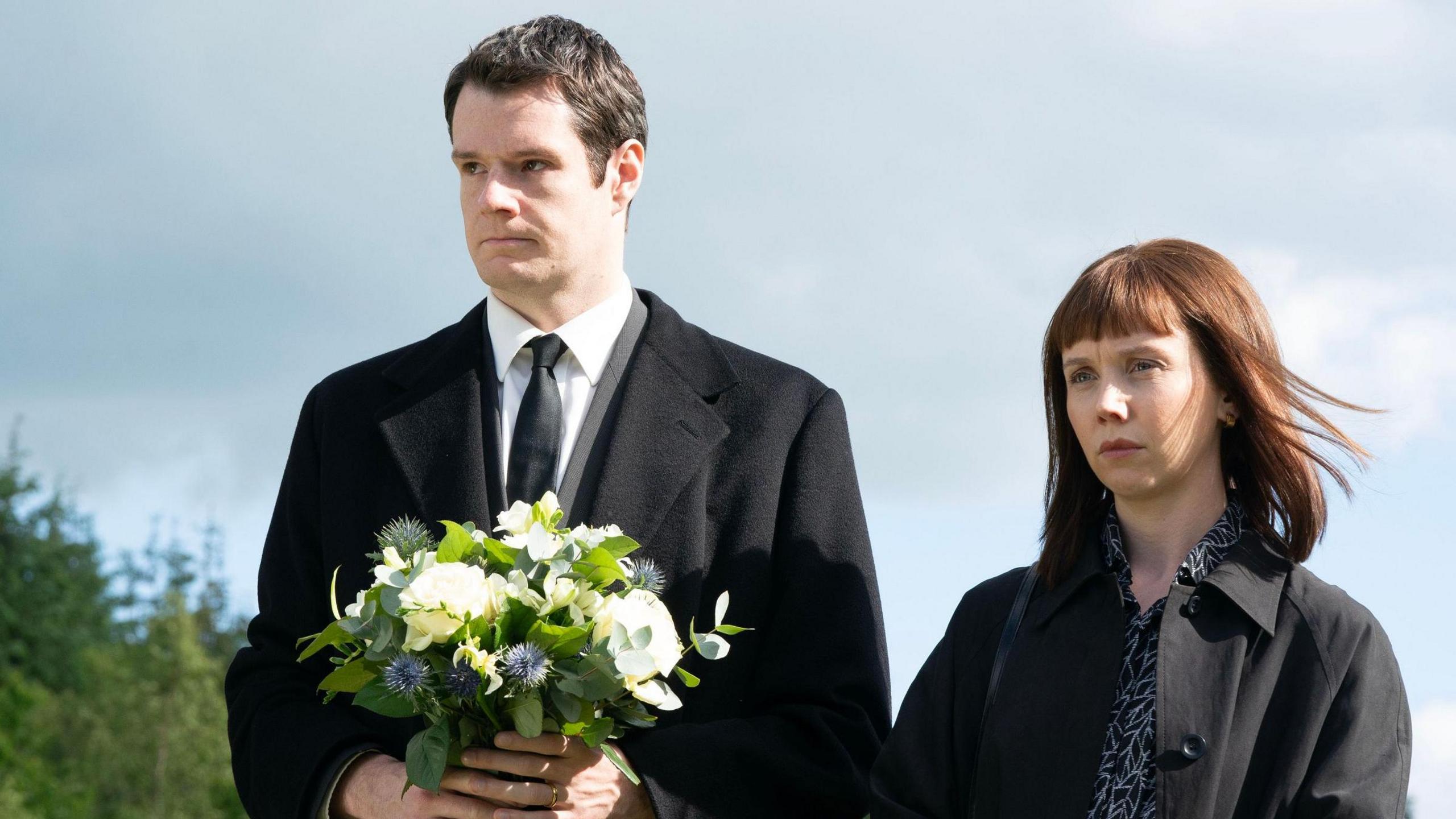 Actors Connor Swindells and Lauren Lyle in dark clothes holding flowers outside, at a memorial