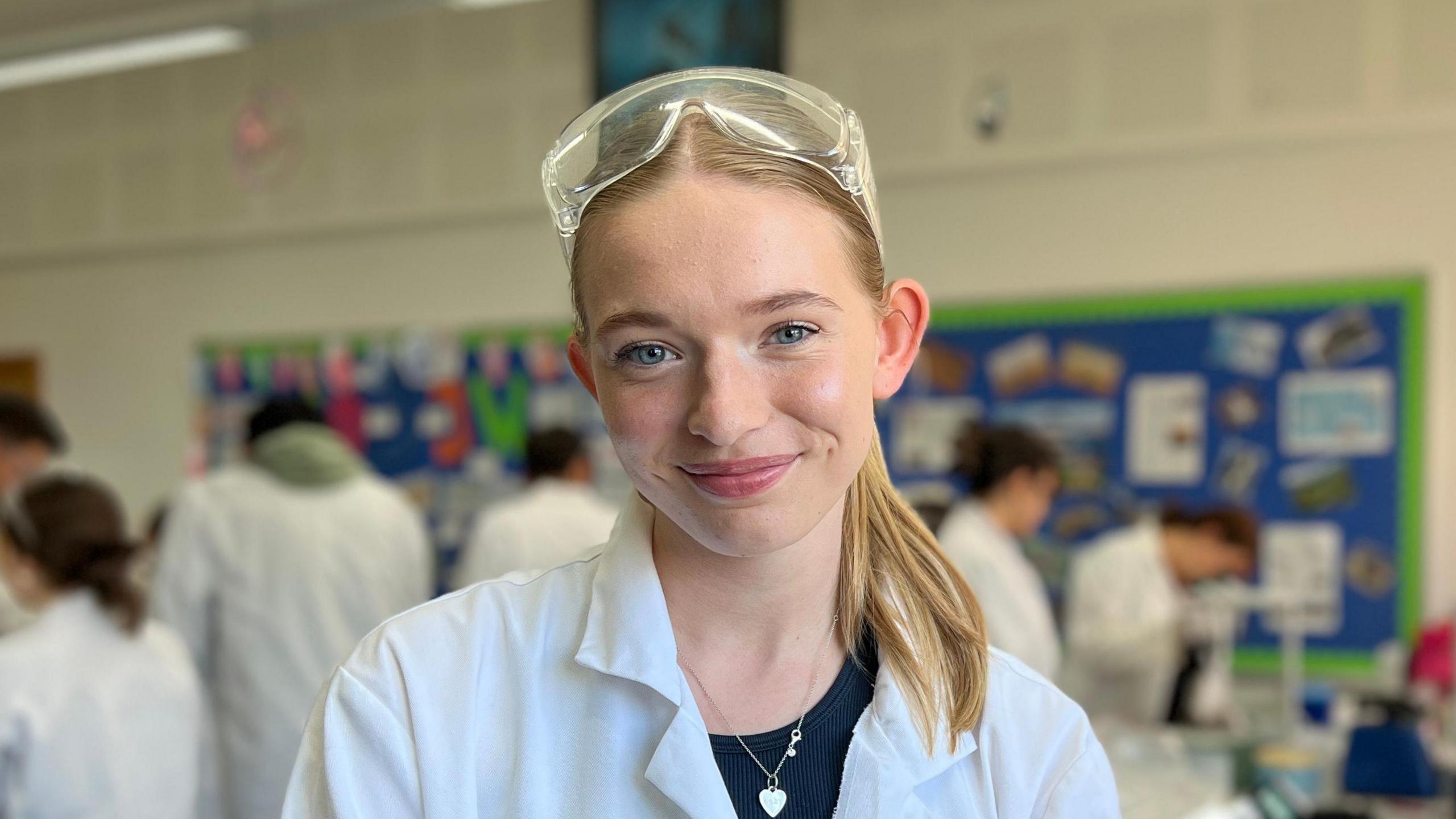 A classroom filled with students wearing white lab coats and safety goggles, engaged in various scientific activities. The focus is on Evie who is standing in the foreground, with other students working at lab benches in the background. The classroom has a bulletin board with colourful posters and a large window letting in natural light.