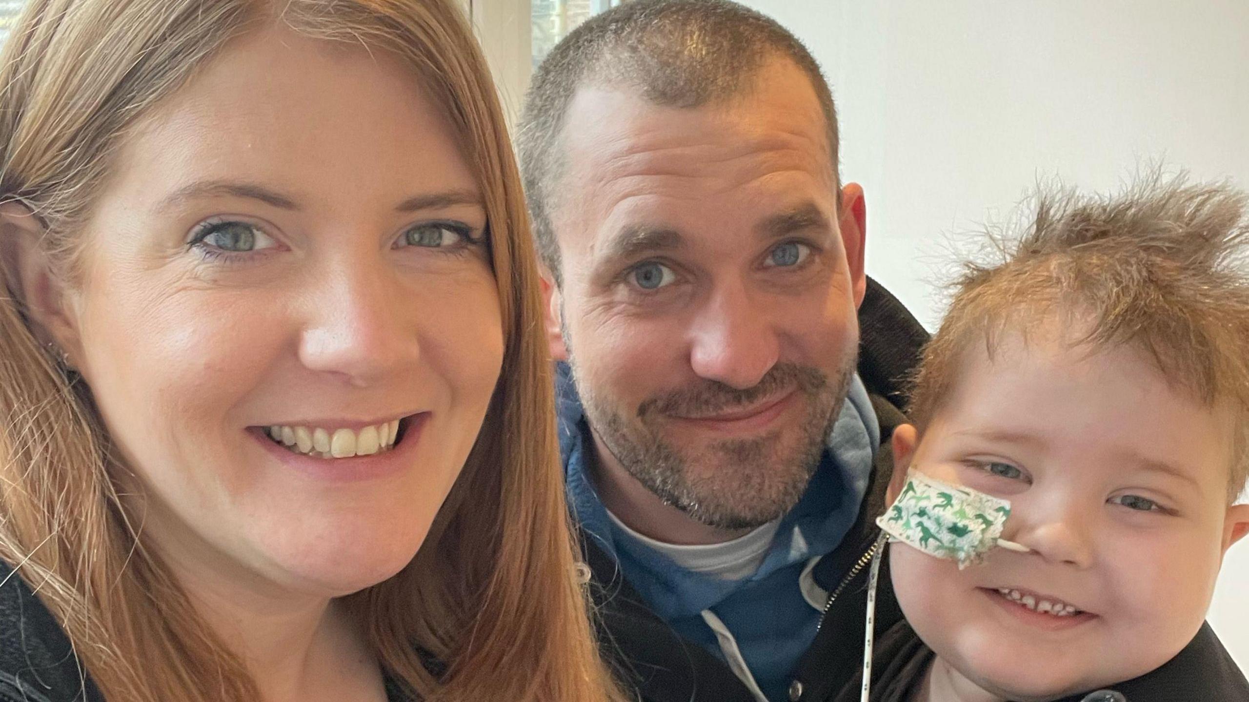 A mum and dad with their young son, who has a medical tube coming from his nose. All are smiling at the camera.