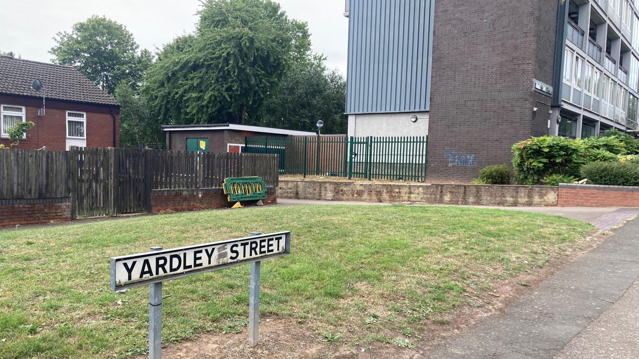 A sign on a green area of grass which says Yardley Street. Some of the grass has brown patches. In the background is a wooden fence and a house and to the right is the bottom of a block of flats. 