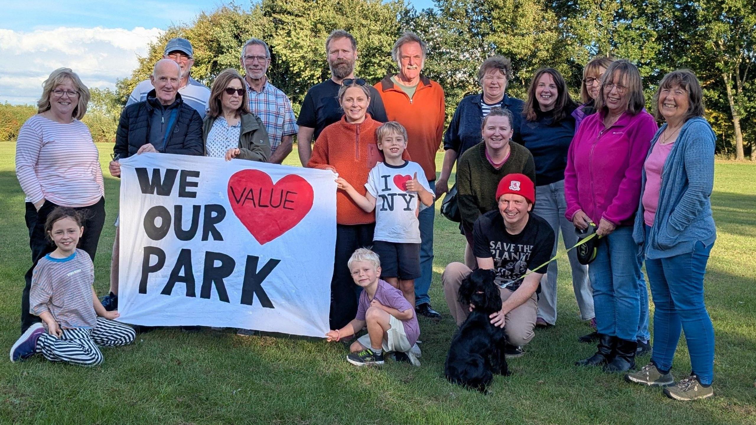 A mixed age group of residents - children and adults - from North Cowton village stand in a open green space. Several people hold up a white sign with black lettering and a red love heart which reads: 'We Value Our Park'.
