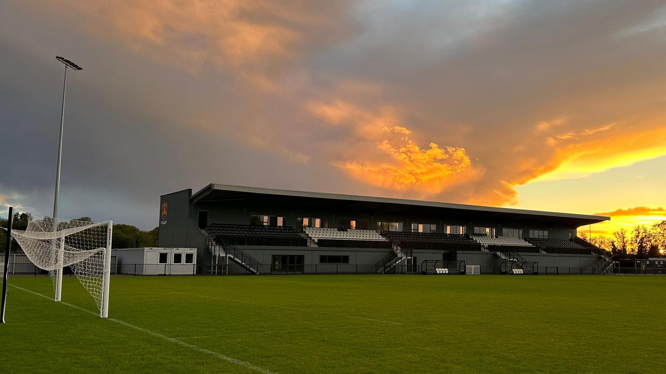 Cambridge City FC ground at Sawston. A grey football stadium stand and the above is a yellow/orange sunset. There is also a goal with the net folded up at the end of the pitch which runs in front of the stand