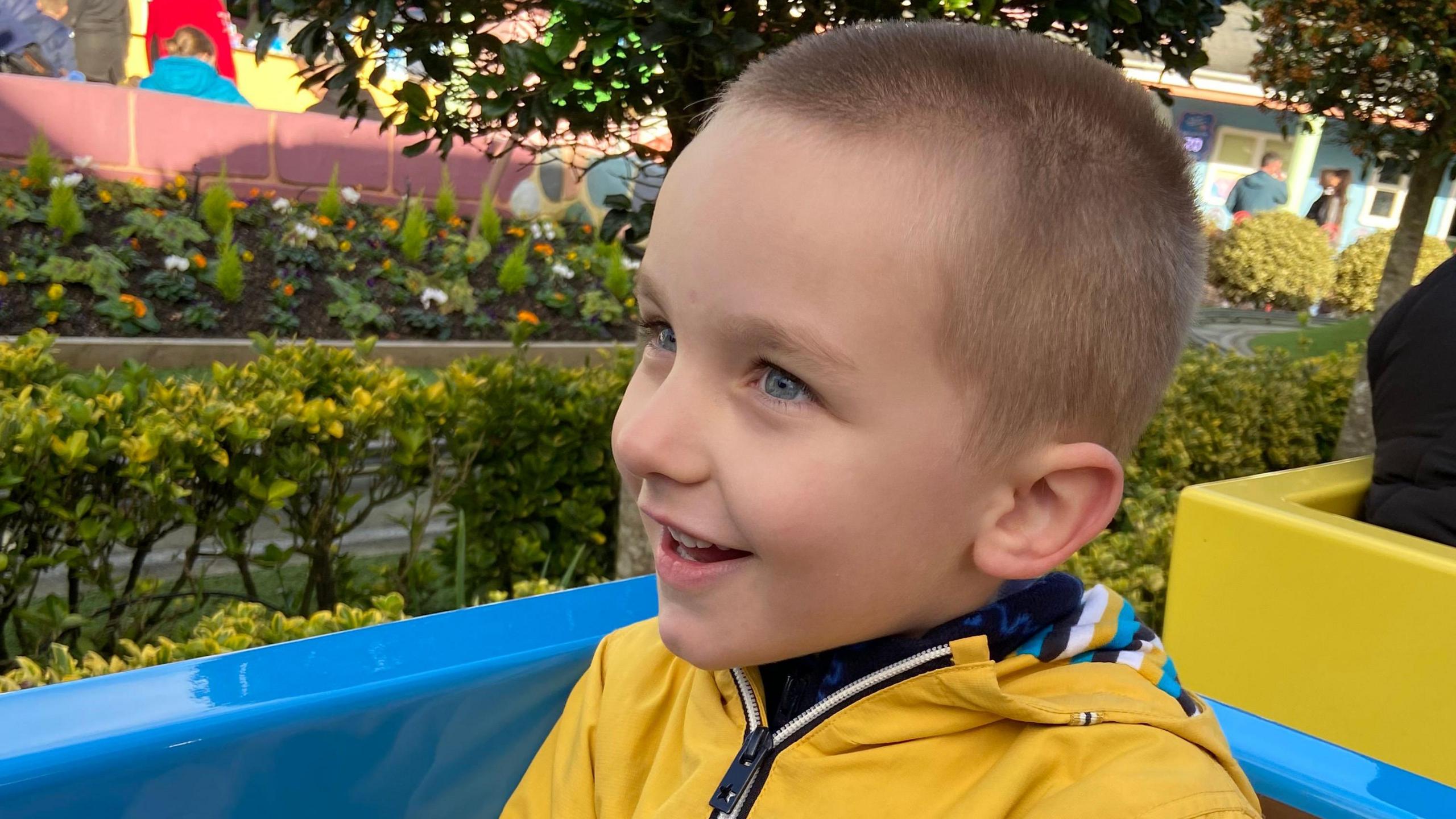 Edward is smiling whilst on a ride. He has short hair and is wearing a yellow coat.