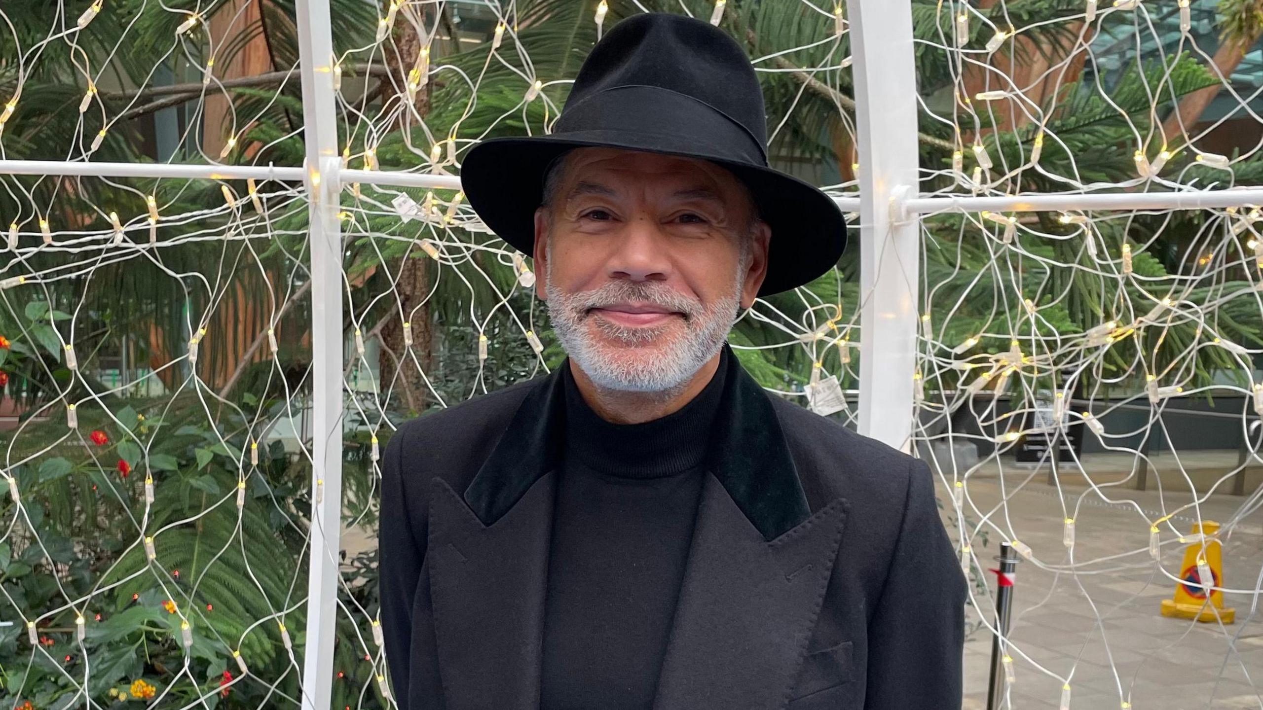 Russell McFarlane, a mixed-race man in his 60s with a short, white beard. He's smiling and wears an all-black suit with a black Panama hat.