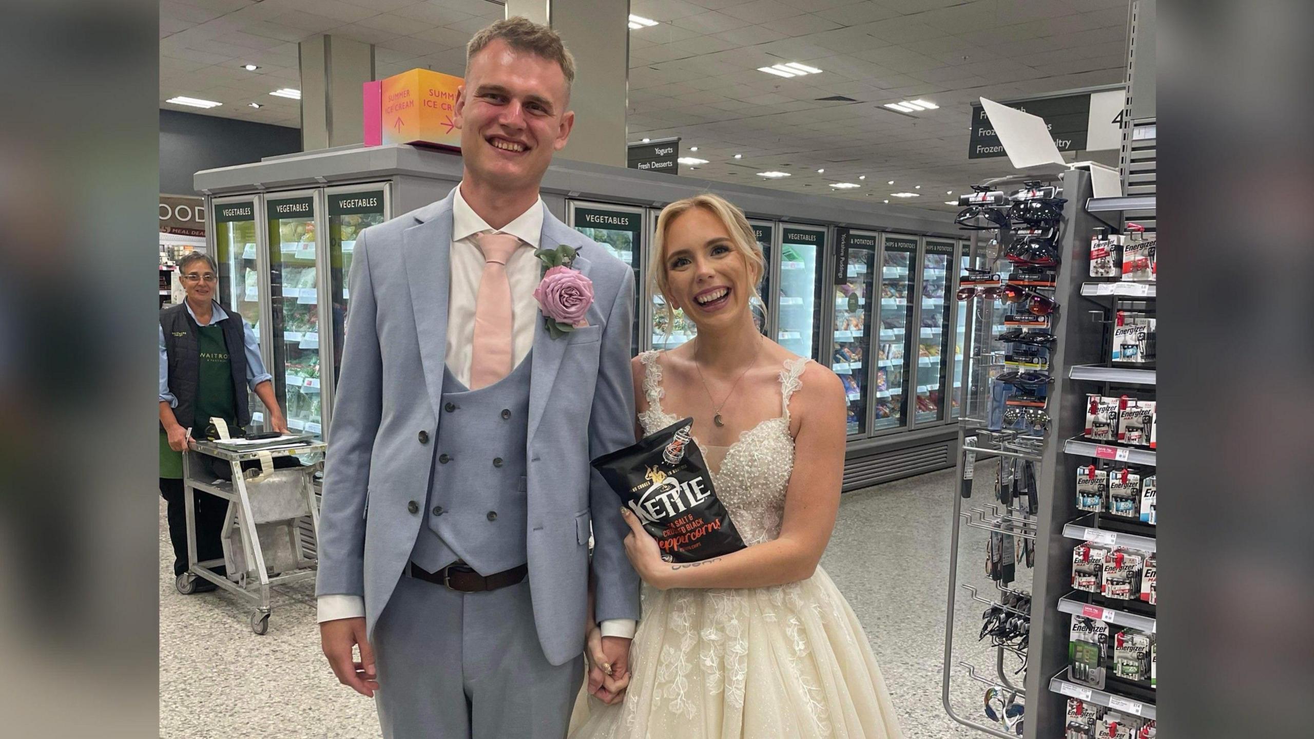 A man in a blue three-piece suit and a woman in a bridal gown hold hands and a packet of crisps in the aisle of a supermarket. 