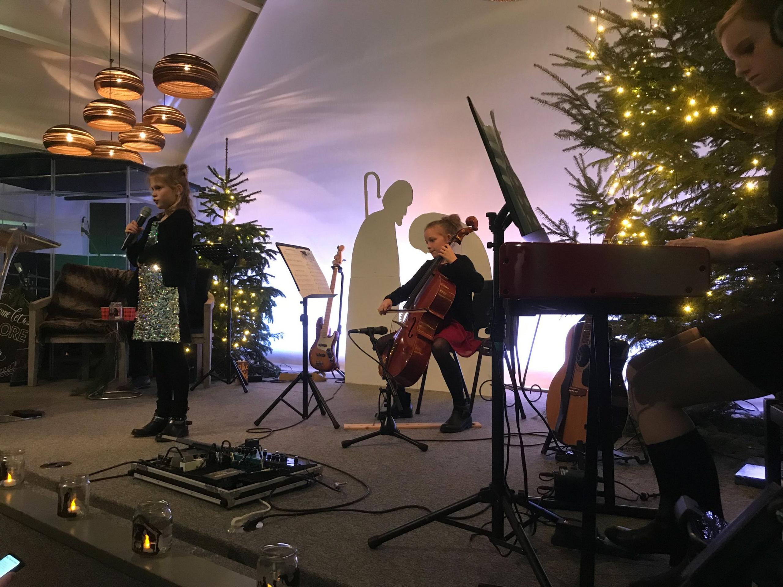 Girl singing at the front of stage during a school performance. Behind her is another girl on the cello and a person playing the piano. The stage is decorated with Christmas trees and there is a guitar on a stand in the background. 