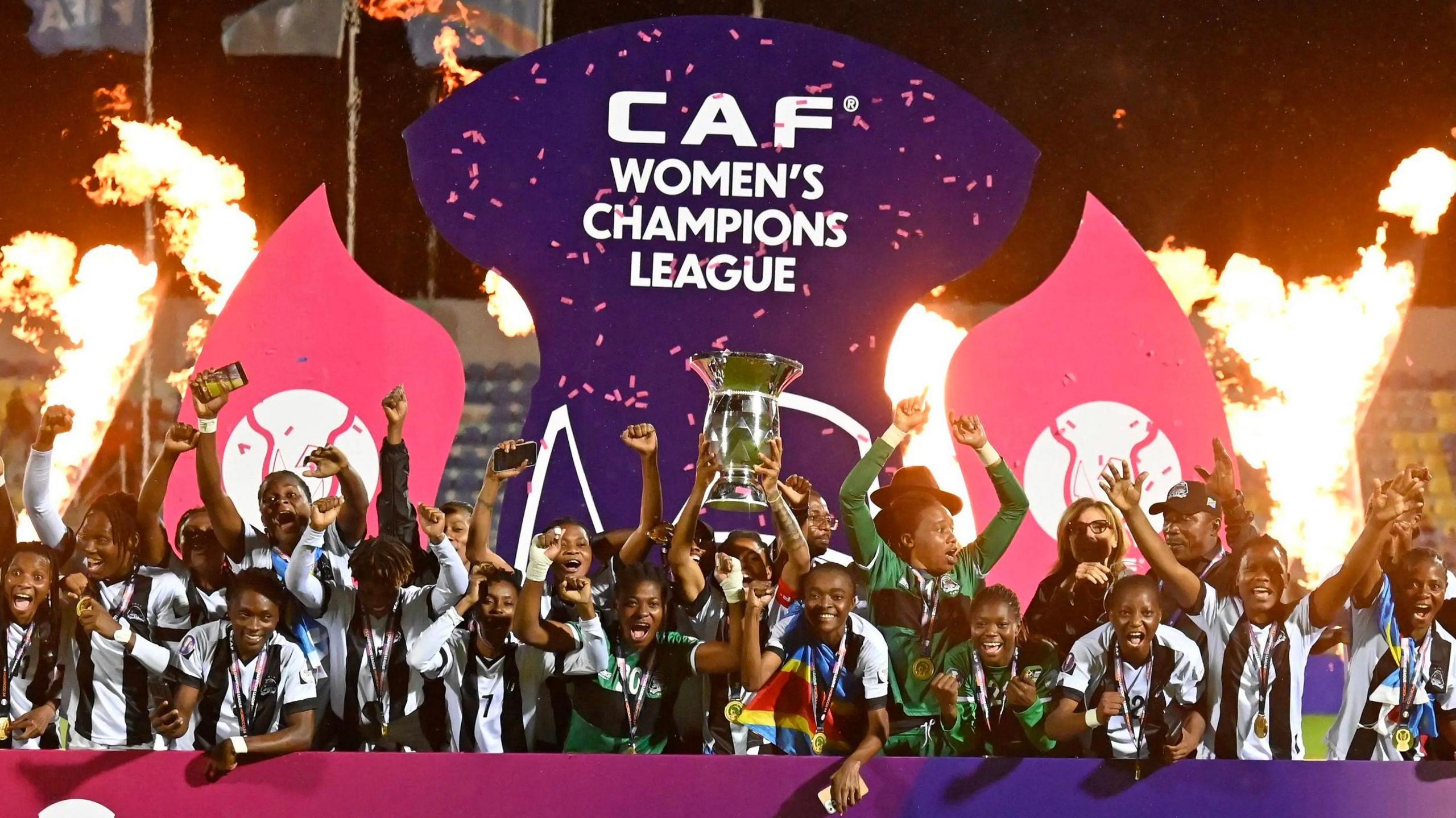 TP Mazembe players, wearing black and white shirts, celebrate as they lift the African Women's Champions League trophy against a pink and purple branded hoarding and bursts of flame in the background