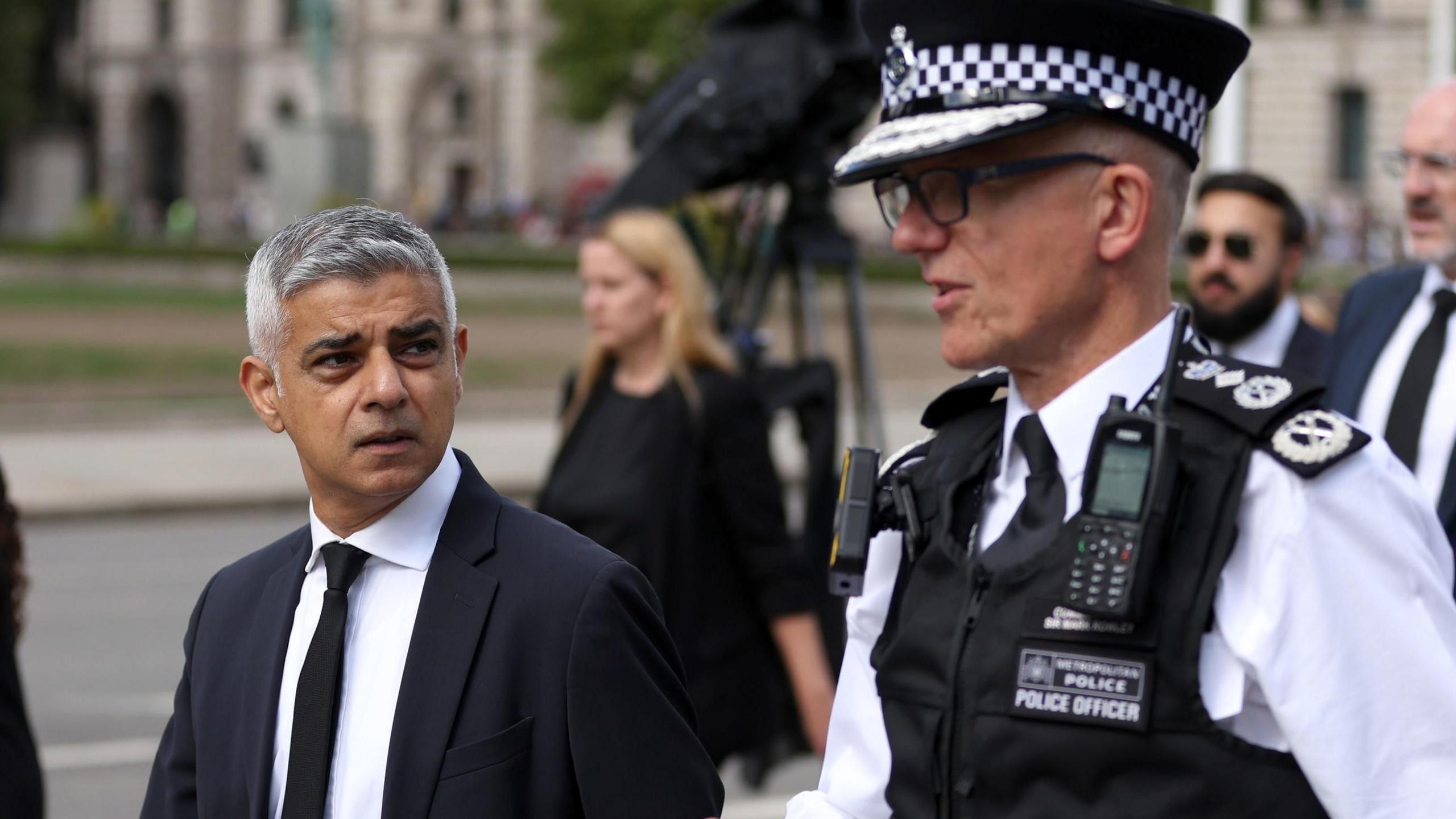 Sadiq Khan walking outside and speaking with a Metropolitan Police commissioner Sir Mark Rowley in uniform, with other people and buildings visible in the background.