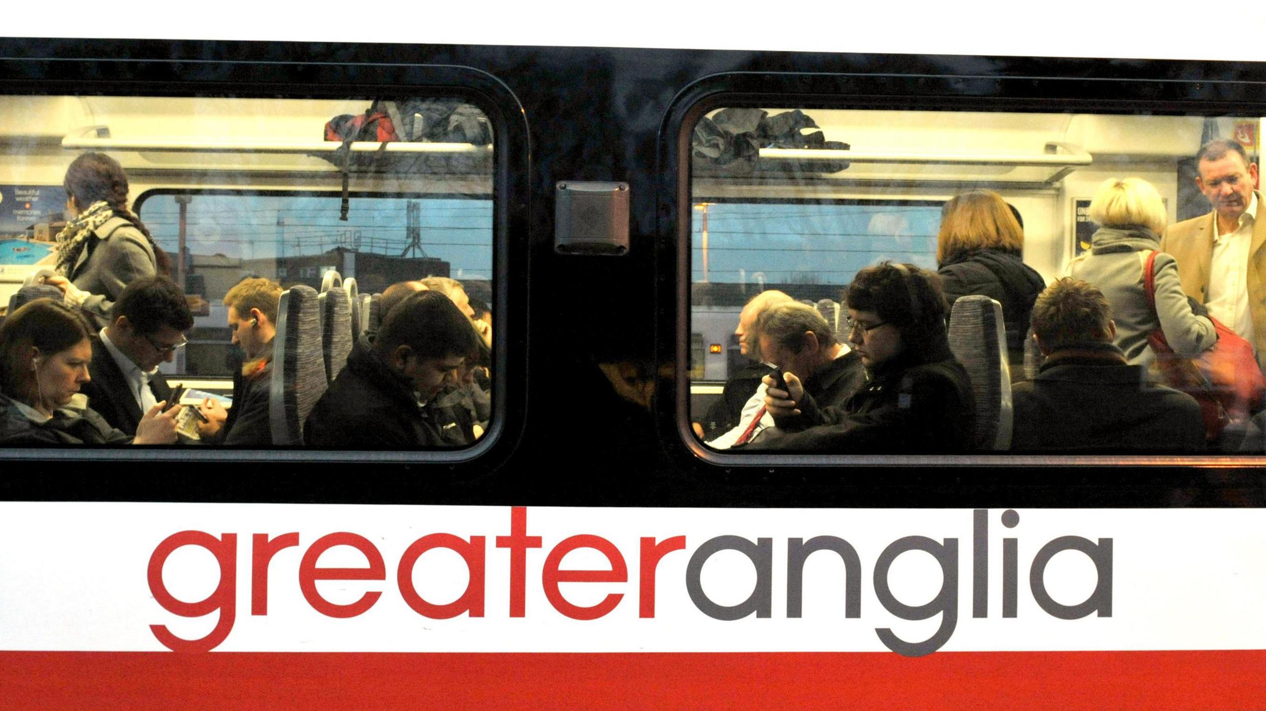 Commuters on a greater Anglia train. The picture has been taken from outside the train  and shows the greater anglia sign on the side below two windows. Passengers can be seen sitting and standing on the train. 