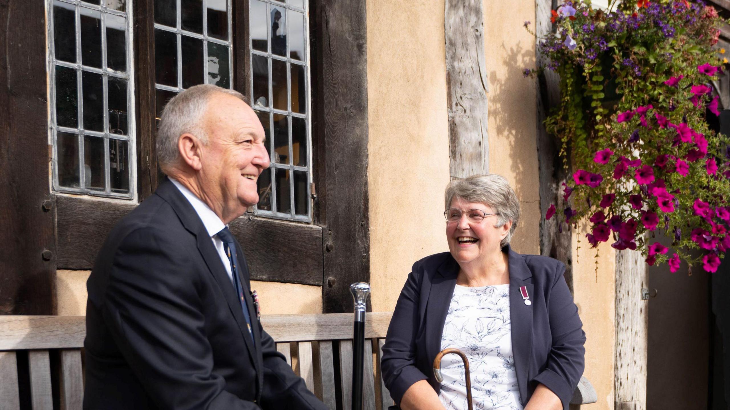 Major (rtd) Brodie-Murphy and Captain (rtd) Gill sat on a bench laughing in front of the Lord Leycester Hospital which is a Tudor-style building painted yellow with exposed wooden beams. 