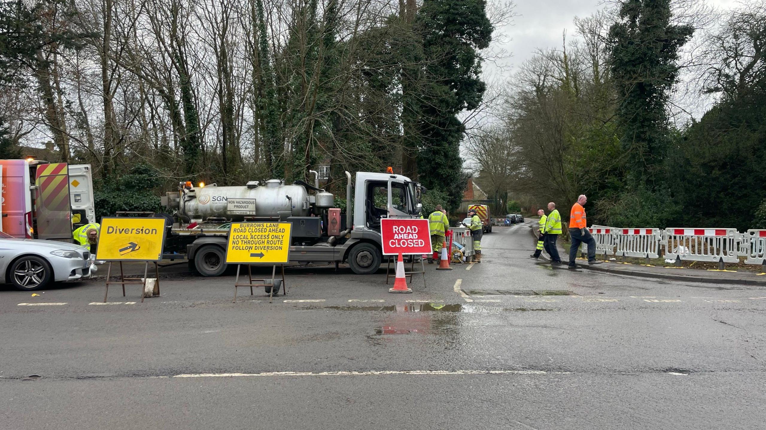 Road signs with a SGN fences blocking the road. There are multiple men wearing high-visibility jackets stood close to a large tanker. 