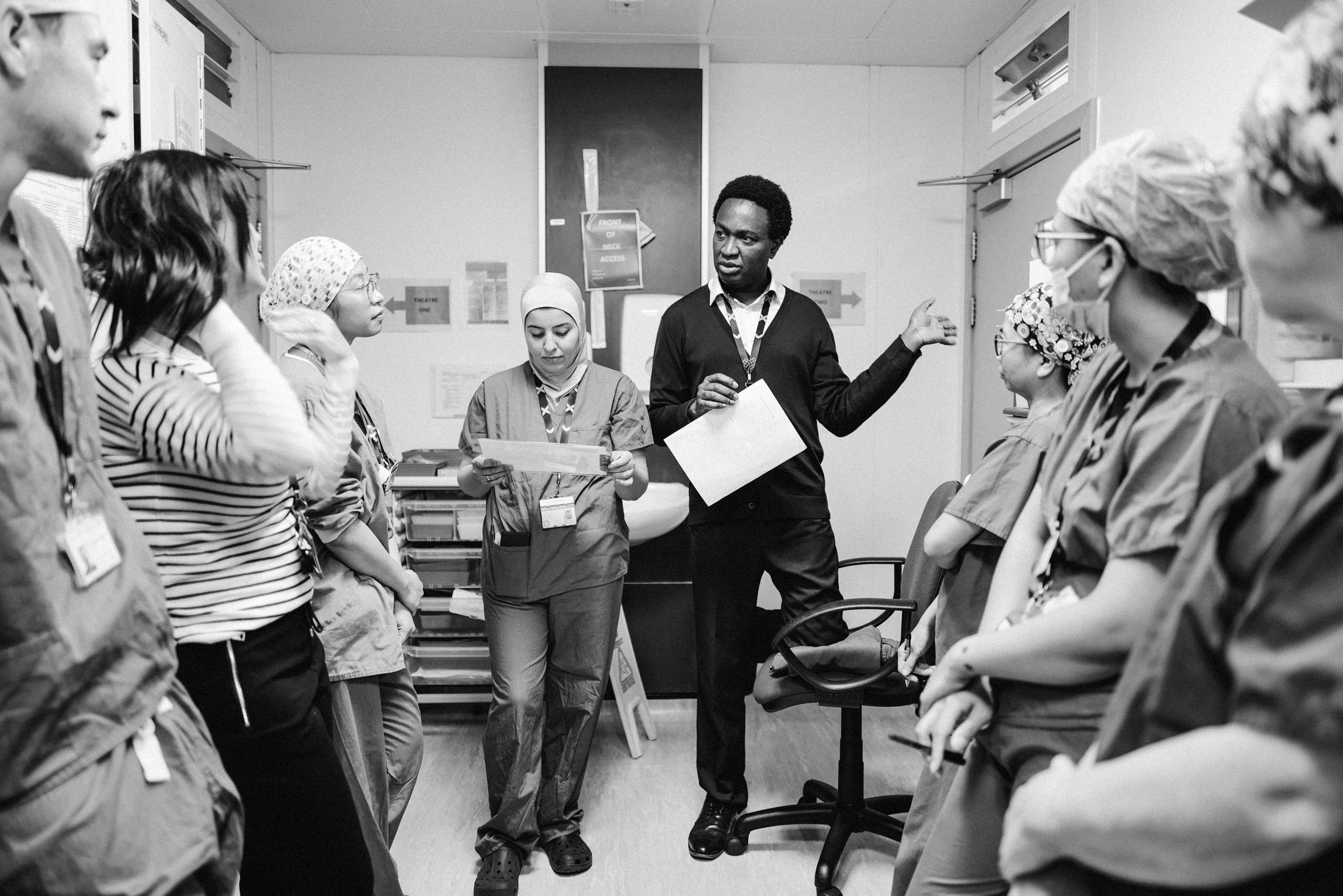 All images in black and white. Eight maternity ward staff, including some in scrubs, are being briefed on the upcoming procedure.