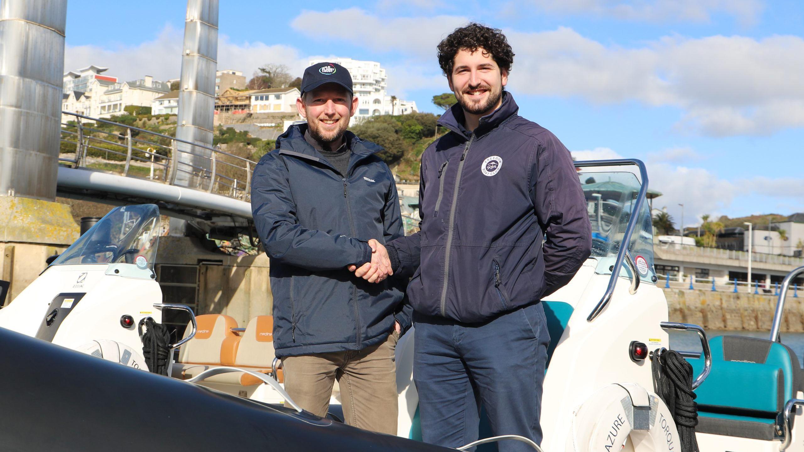 Dr Andrew Bowkett for Wild Planet Trust (left) shakes hands with Oliver Patterson from Freedom Boat Club Torquay on a boat. It is a sunny day with a few clouds in the sky. Both mean are wearing dark blue coats. Dr Bowkett has brown chino trousers on. Oliver has navy blue trousers. Both men have goatee beards.