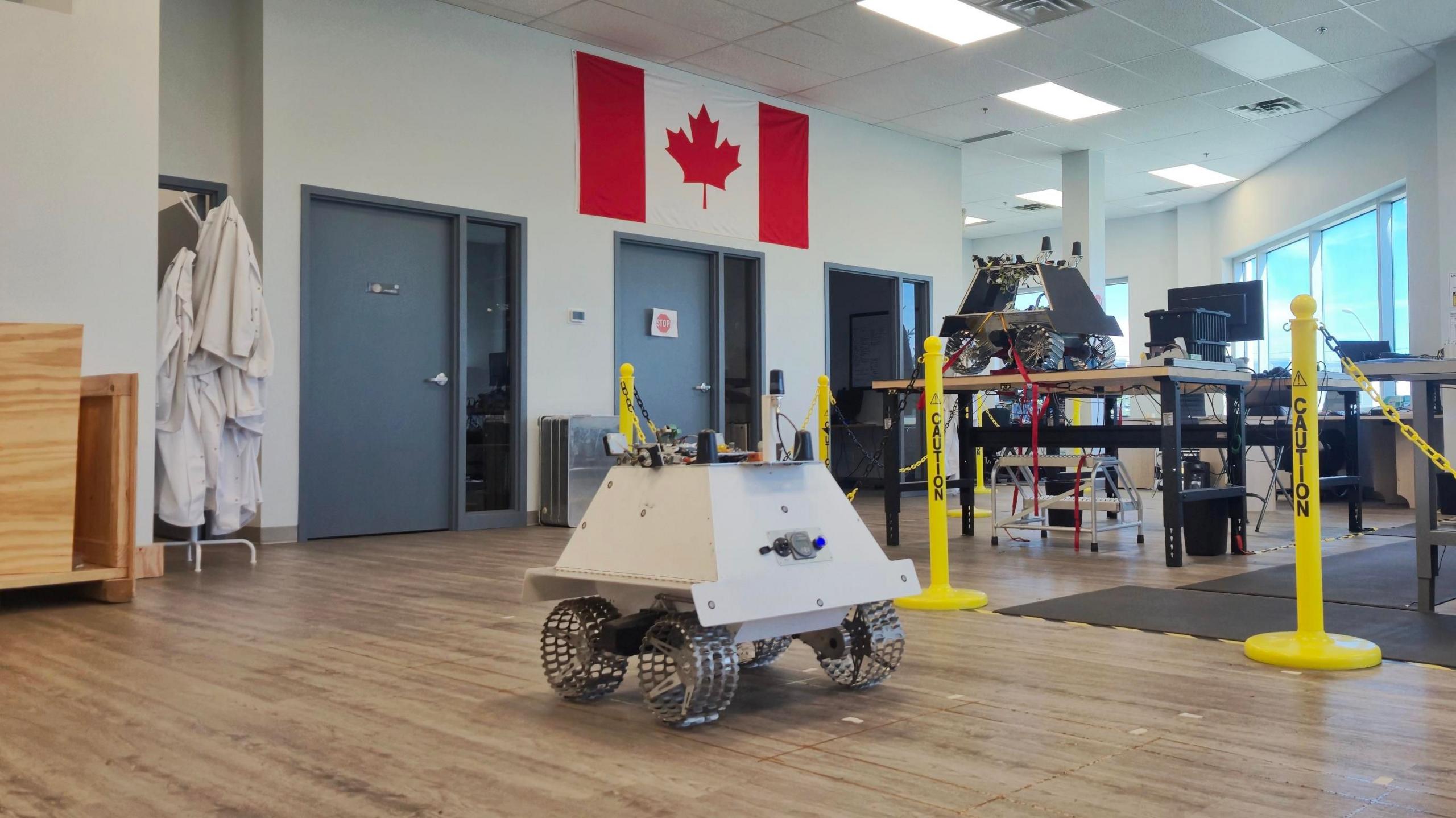 Two prototypes of the Moon rover, one white one in the foreground and the grey one on the table, with the Canadian flag in the background