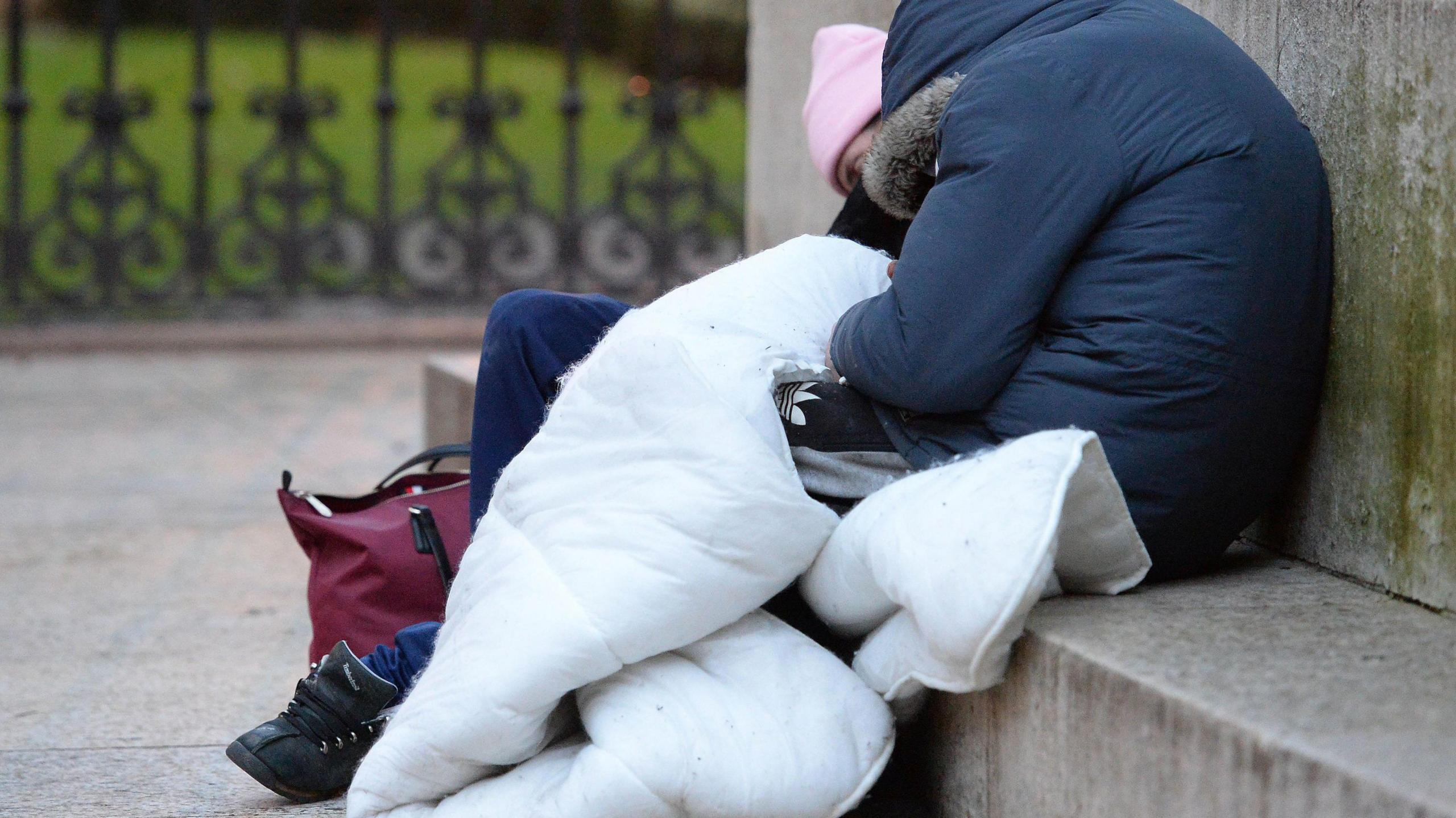 A person wrapped in a duvet is falling asleep on some stone steps in a park. They are wearing a padded dark blue jacket and a pink woolly hat.