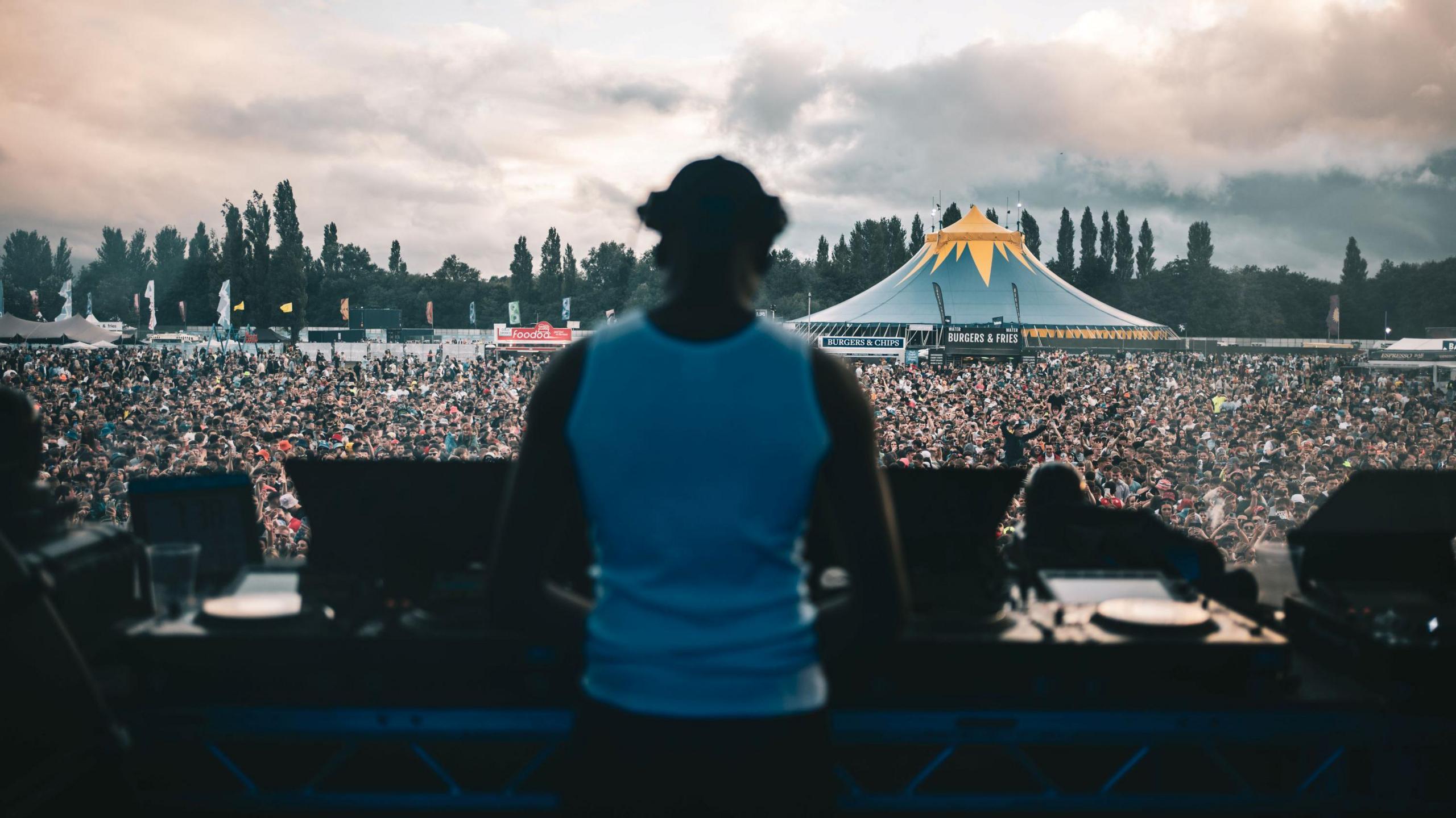 The photo is taken from behind a DJ performing at Emerge. There is a huge crowd of people in front of the stage and one of the tents can be seen in the background.