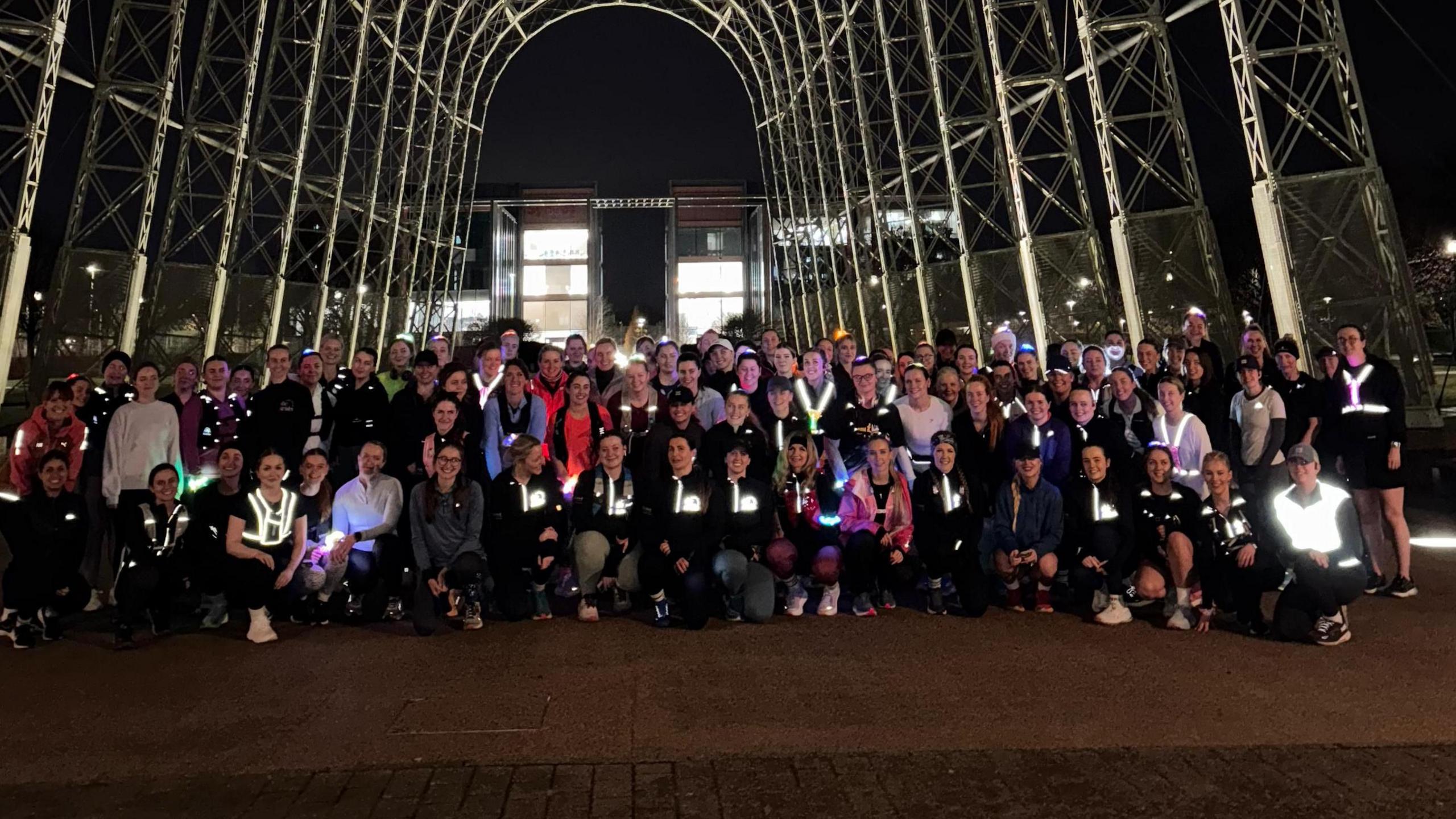 A large group of women pose together at night. They are wearing running gear and many of them have reflective tops. They are underneath a large metal arch and there is an illuminated building behind. 