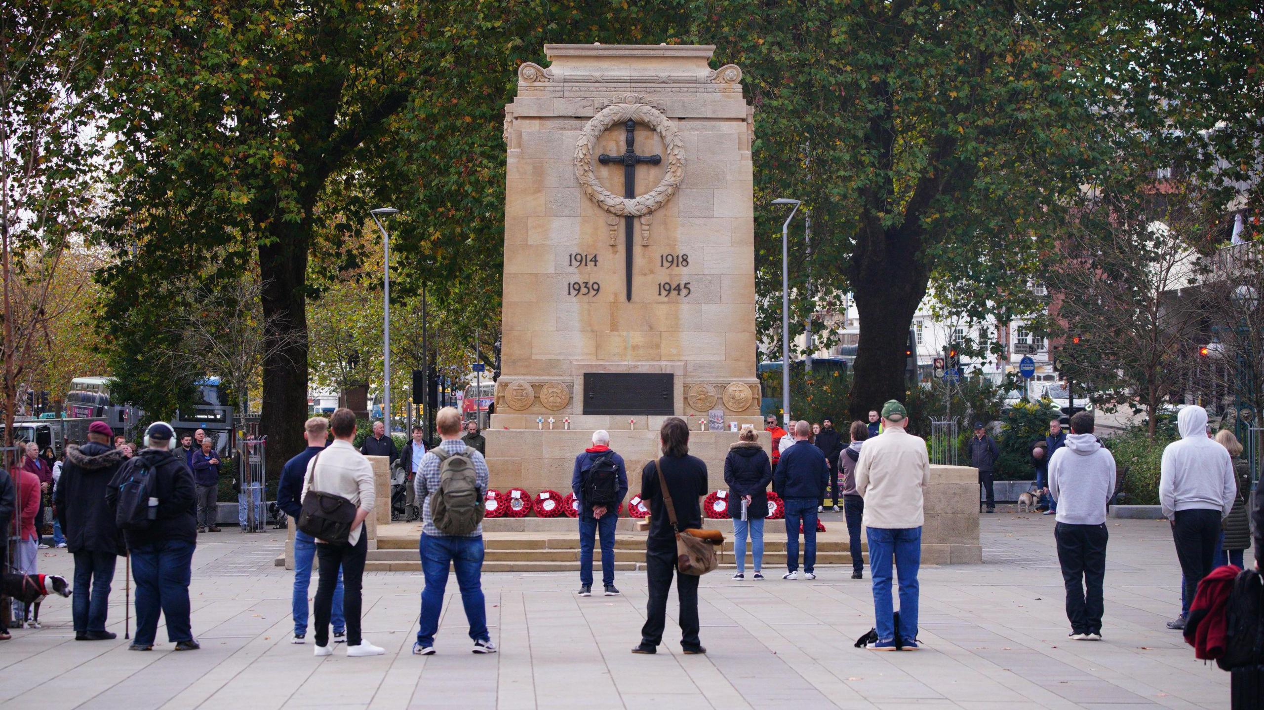 Several people stand still, facing the cenotaph in Bristol city centre, to observe a two minutes' silence for Armistice Day. There are wreaths of poppies at the base of the cenotaph