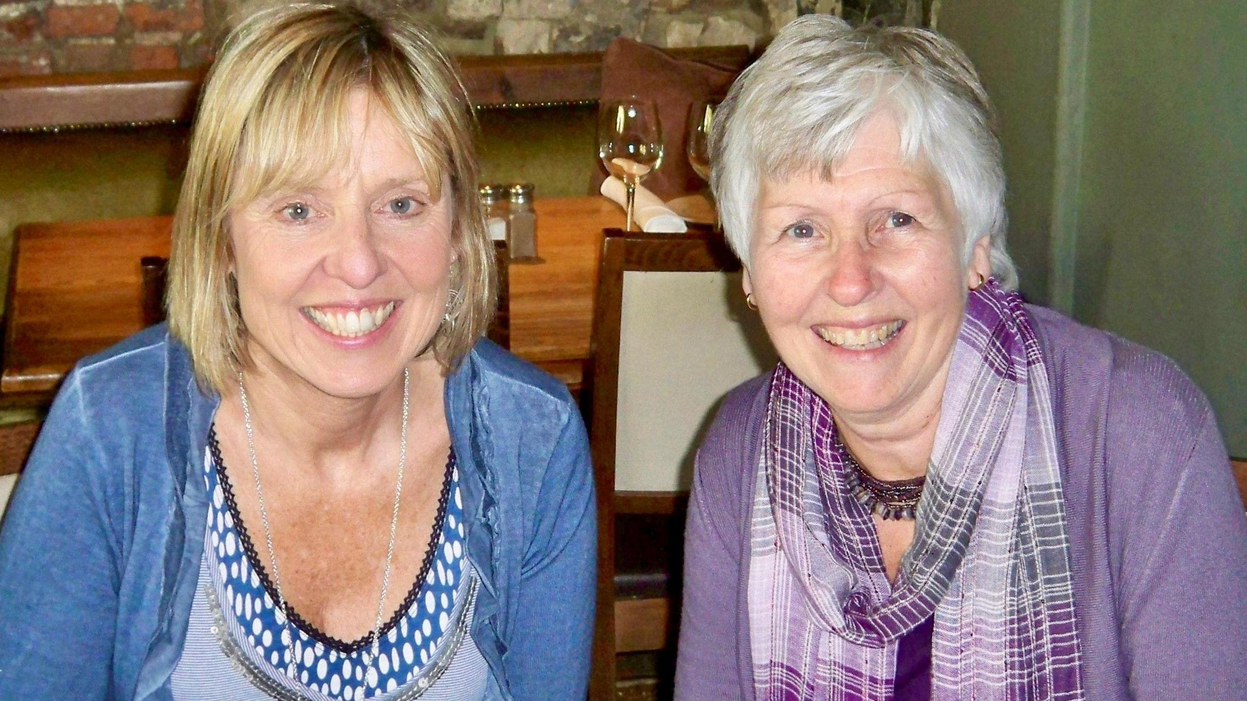 Two ladies who share very similar facial features sit at a table in a restaurant together. The lady on the left has blonde hair and is wearing a blue scoop necked top and blue cardigan. The lady on the right has grey hair and is wearing a purple cardigan with a scarf in a variety of purple and grey shades. 