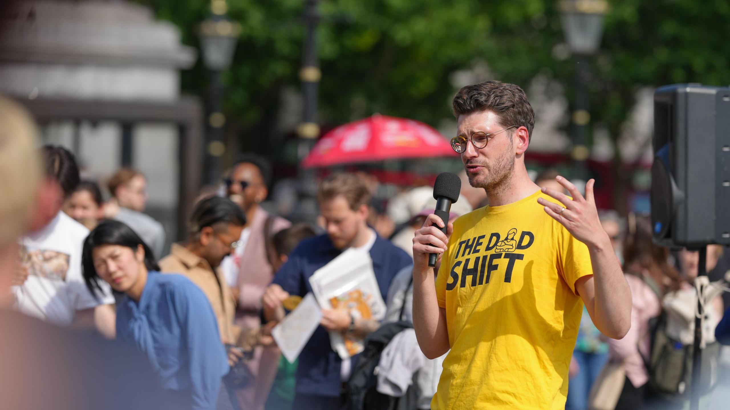 Alex Lloyd Hunter stood in a yellow The Dad Shift t-shirt speaking to a crowd of people