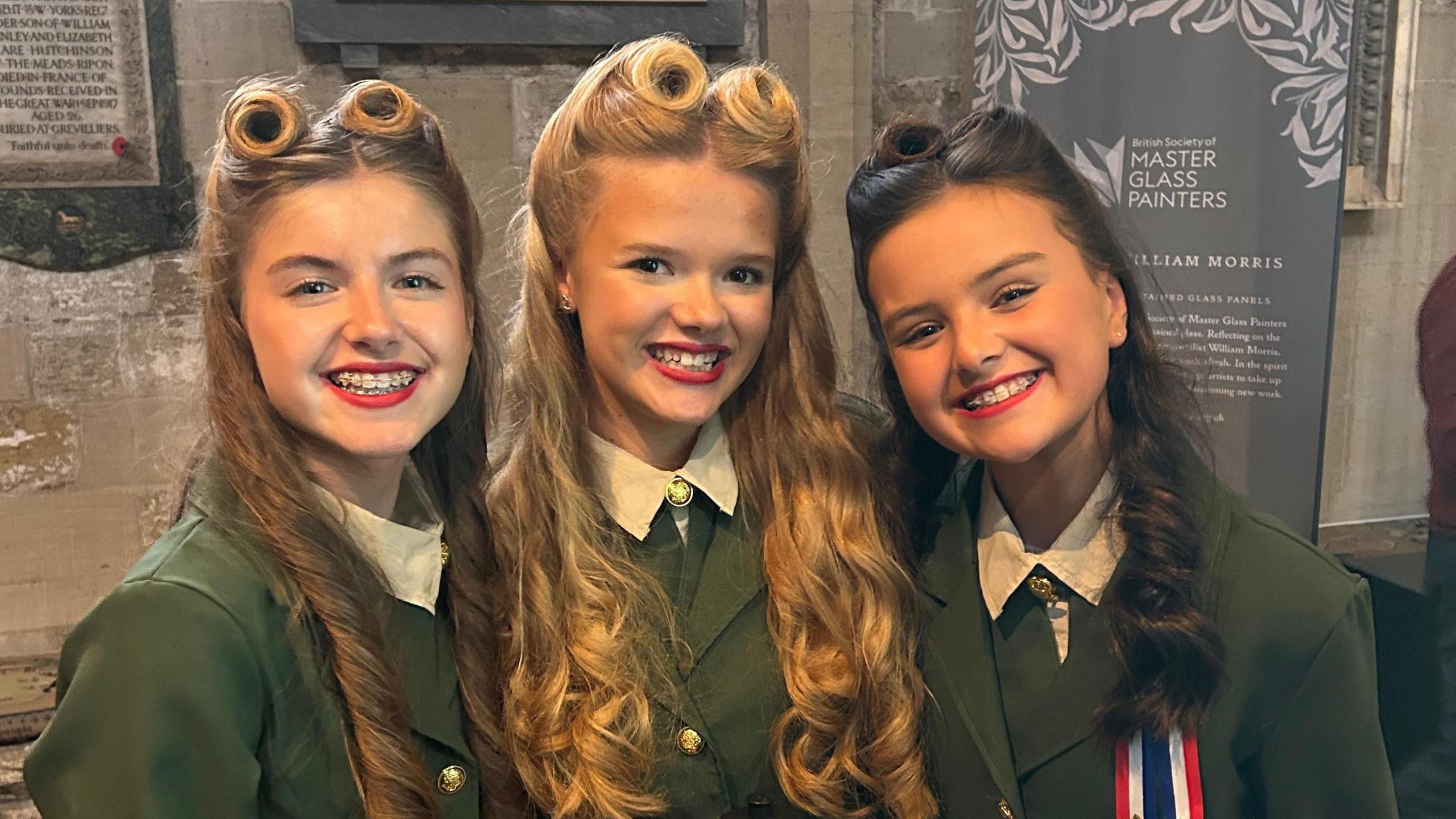 Siena, Emilia and Lottie smile at the camera, against the background of a church. All three girls have long hair with 1940s-style victory rolls, and wear red lipstick and military-style wartime outfits.