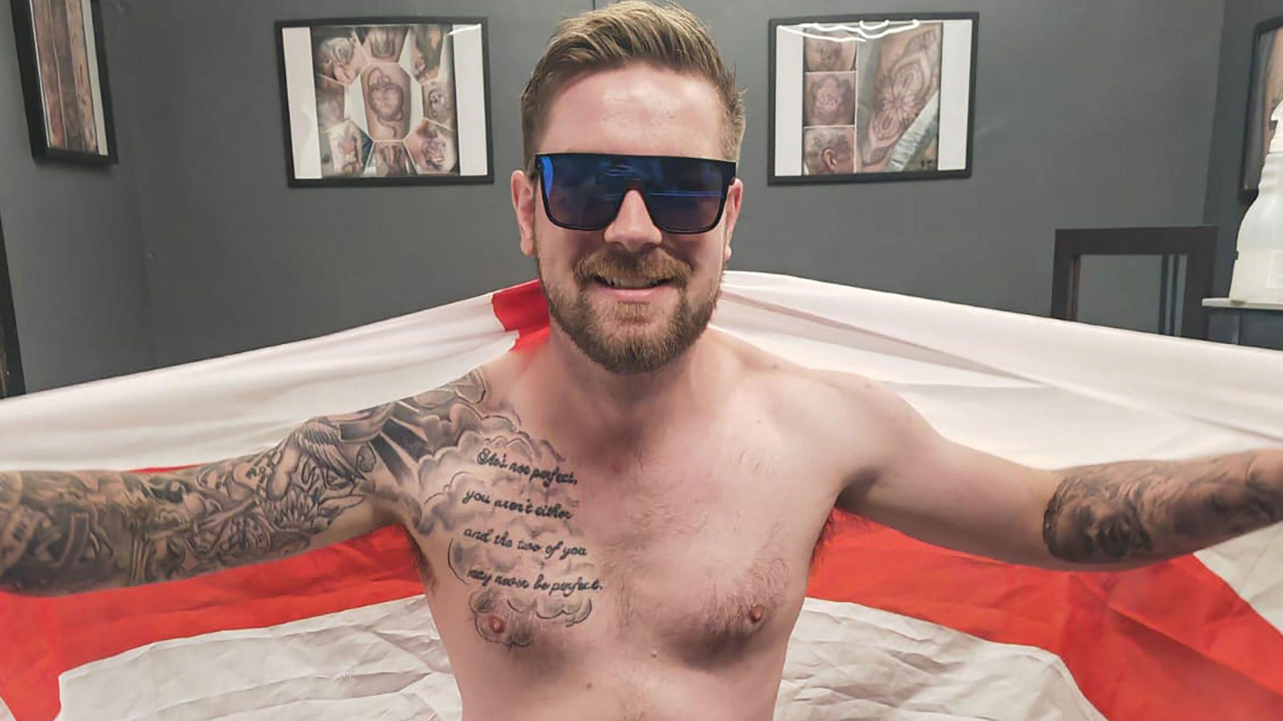 Dan Thomas showing the tattoo of his leg in his pants, holding an England flag