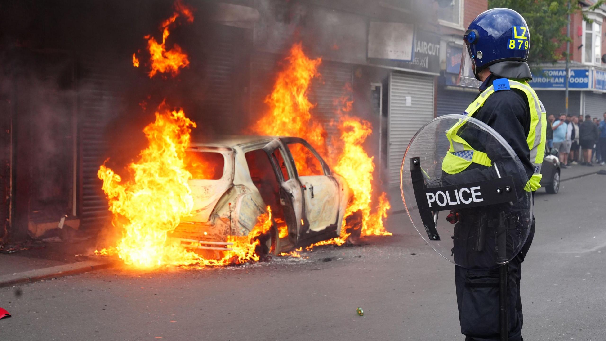 A police officer with a transparent circular shield and in a helmet watches as a car is blazing on fire, with a row of shuttered shops behind.