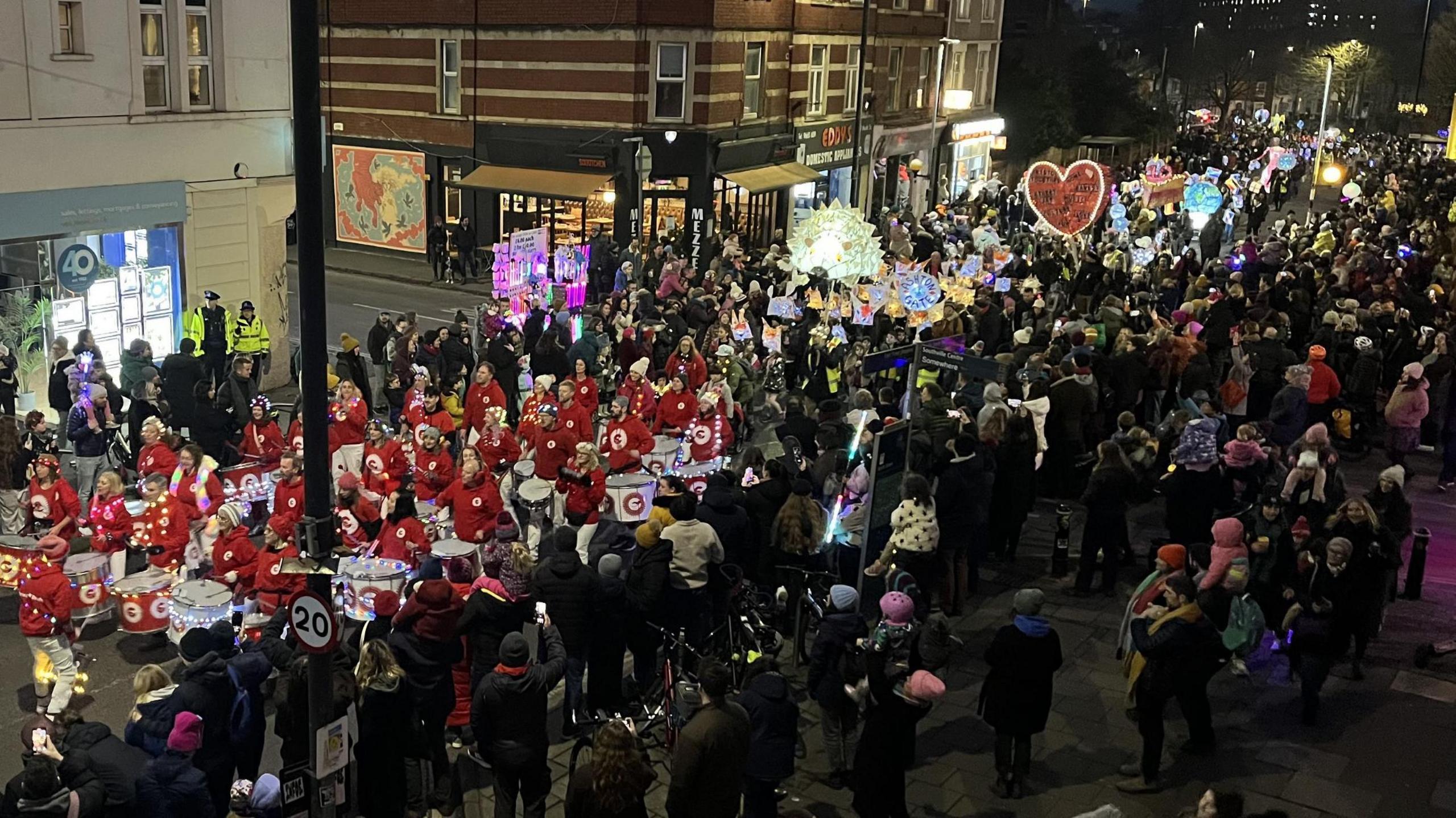 In a picture taken from roof level, North Street in Bedminster in Bristol is crowded with spectators and participants in the annual winter lantern parade. At the front of the procession is a samba band all wearing white trousers and red tops, while behind them many different shaped illuminated lanterns are being carried. The picture is taken at night and shop and restaurant lights are visible along the side of the street