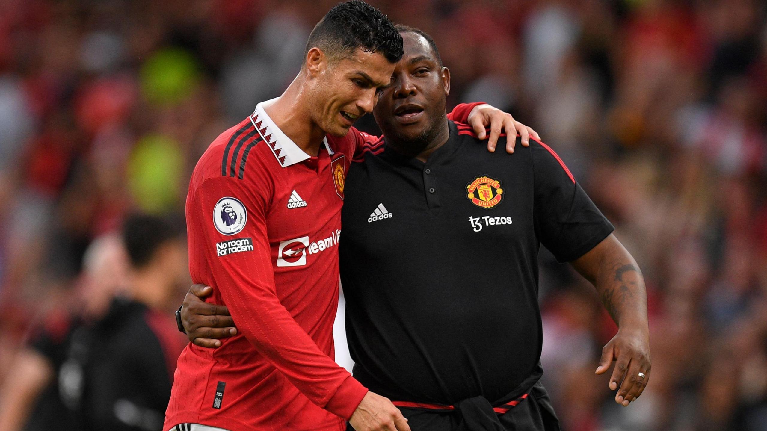 Benni McCarthy, wearing a black Manchester United training top, walks alongside Cristiano Ronaldo, wearing red United kit, with arms wrapped around each other.