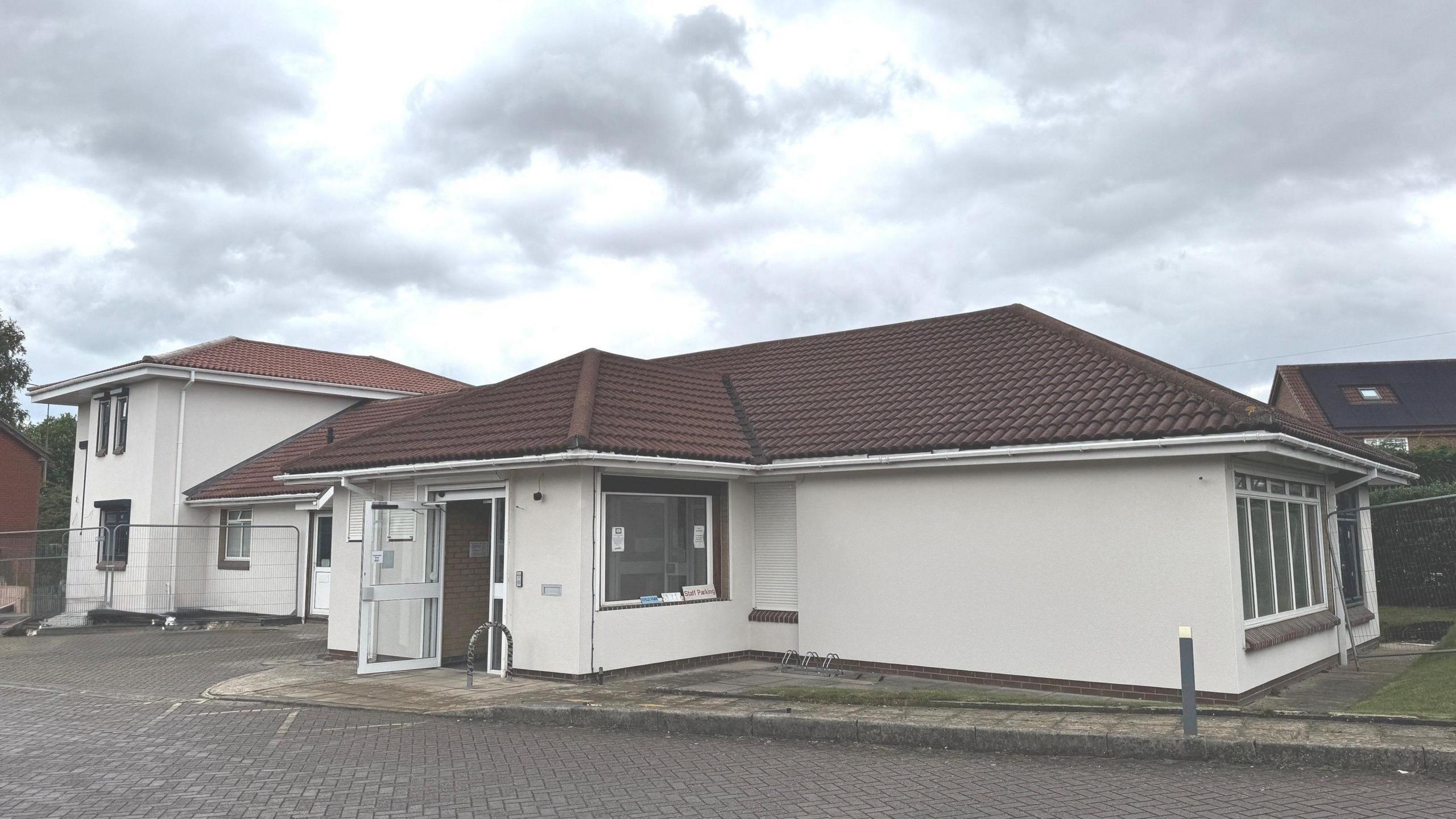 A white rendered single storey building with a brown tiled roof. A white door is open at the front with a path leading to a brick paved road.
