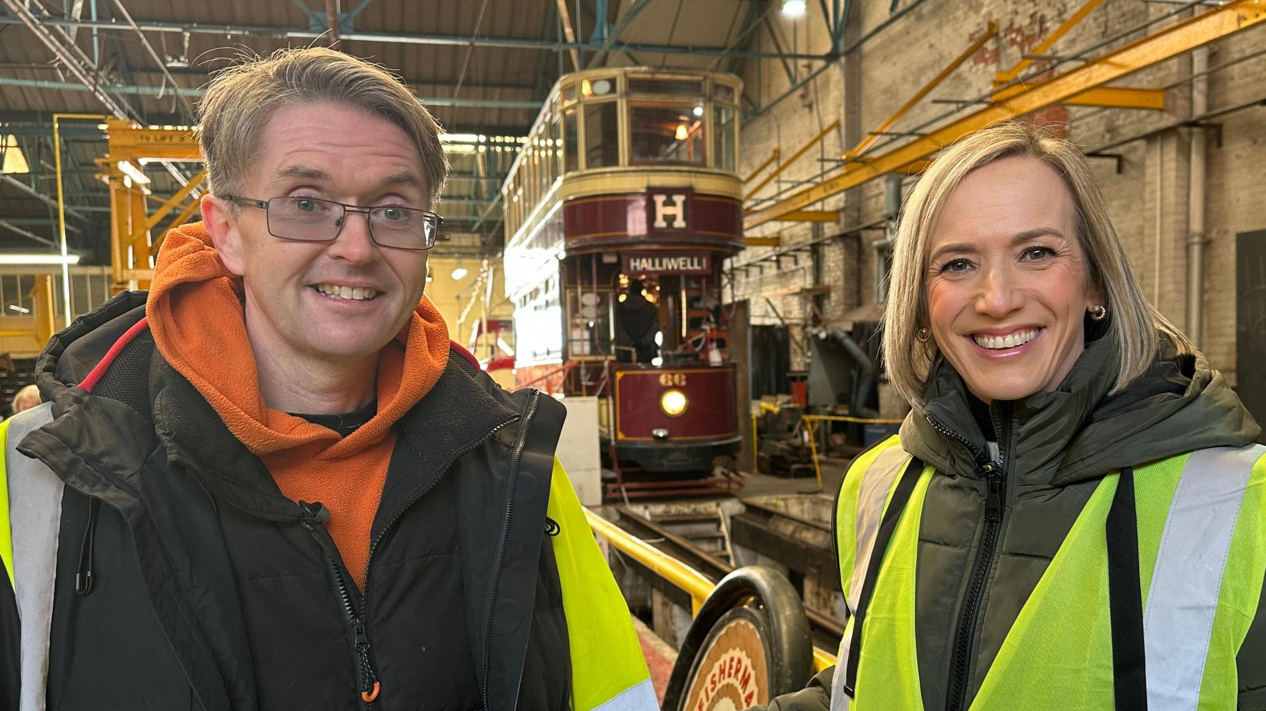 Paul Galley and Kay Crewdson, who are both wearing high-vis jackets, are standing side by side in Tramtown. There are standing in a large warehouse with a maroon-coloured double decker tram in the background. 