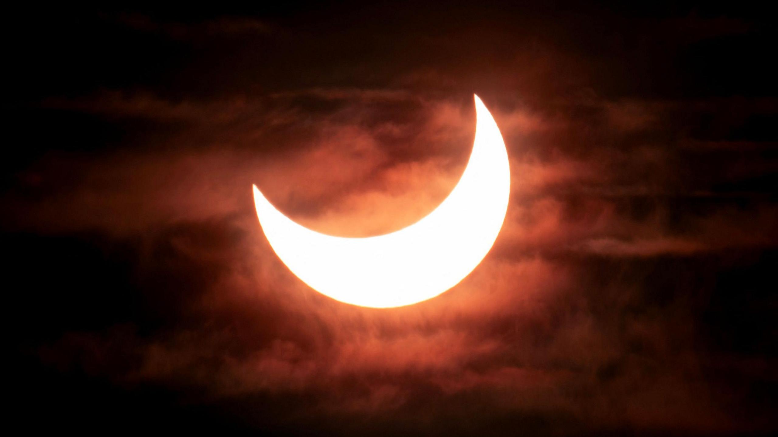 A partial solar eclipse showing the bright crescent of the sun as the moon moves across it from the top left corner. The sky is dark with clouds visible behind the sun