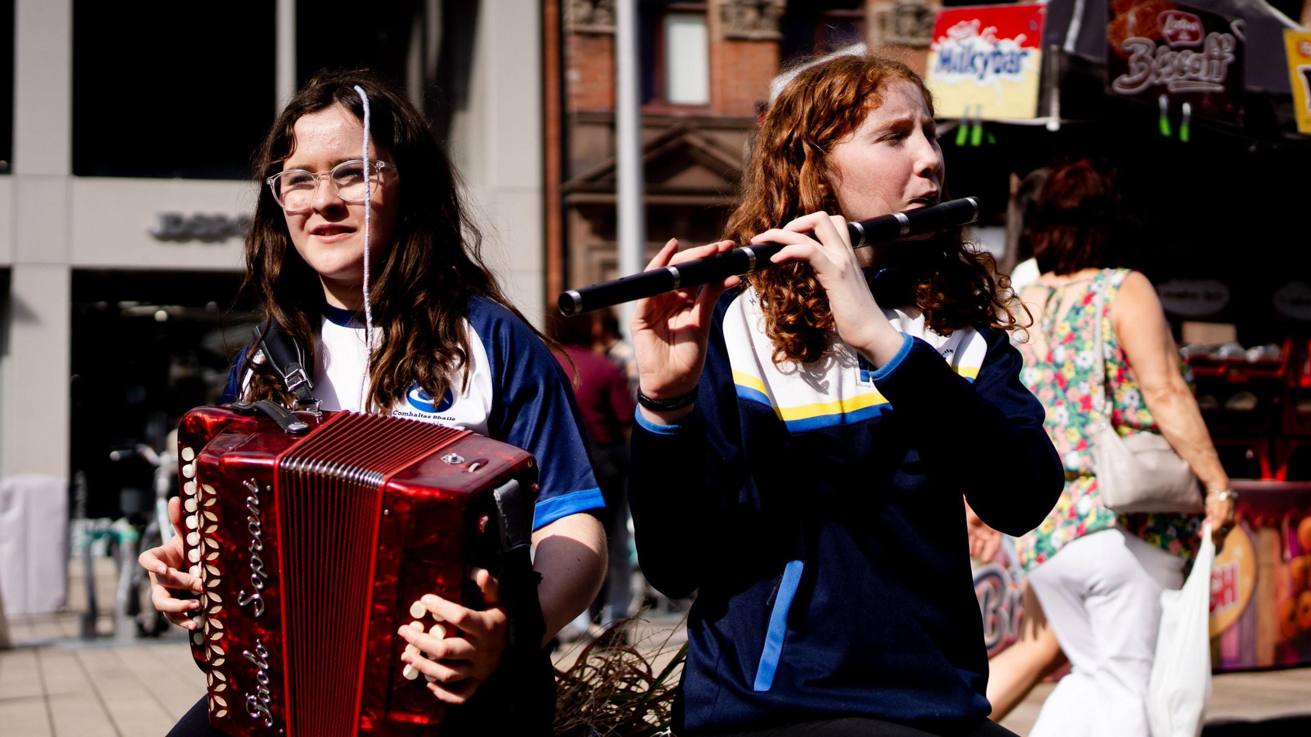 Two young women. One is playing an accordian, the other is playing a flute.